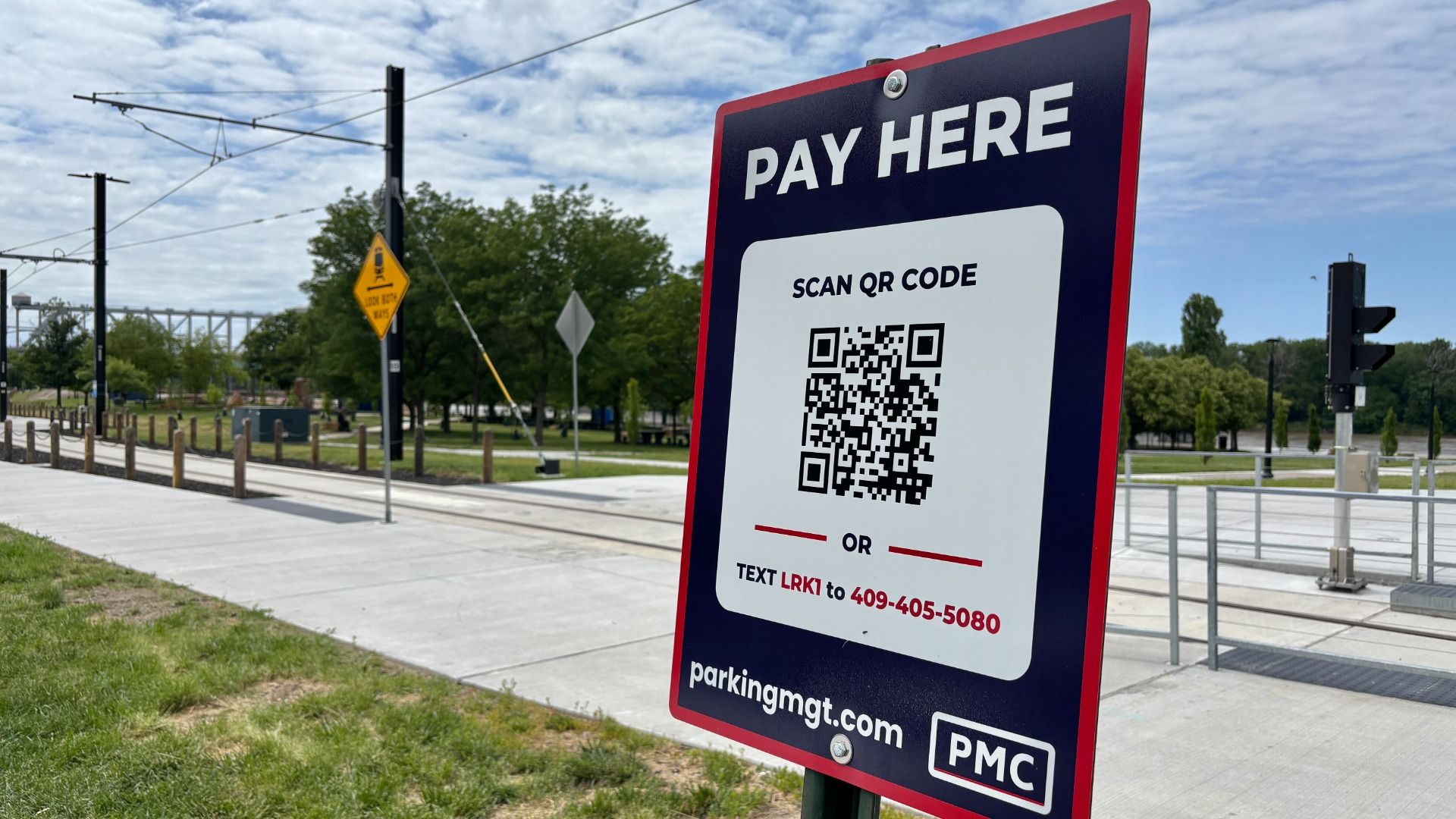 Large blue and red pay here sign with a QR code and instructions to scan or text LRKI; located near railway tracks with overhead wires, trees, and a blue sky in the background.
