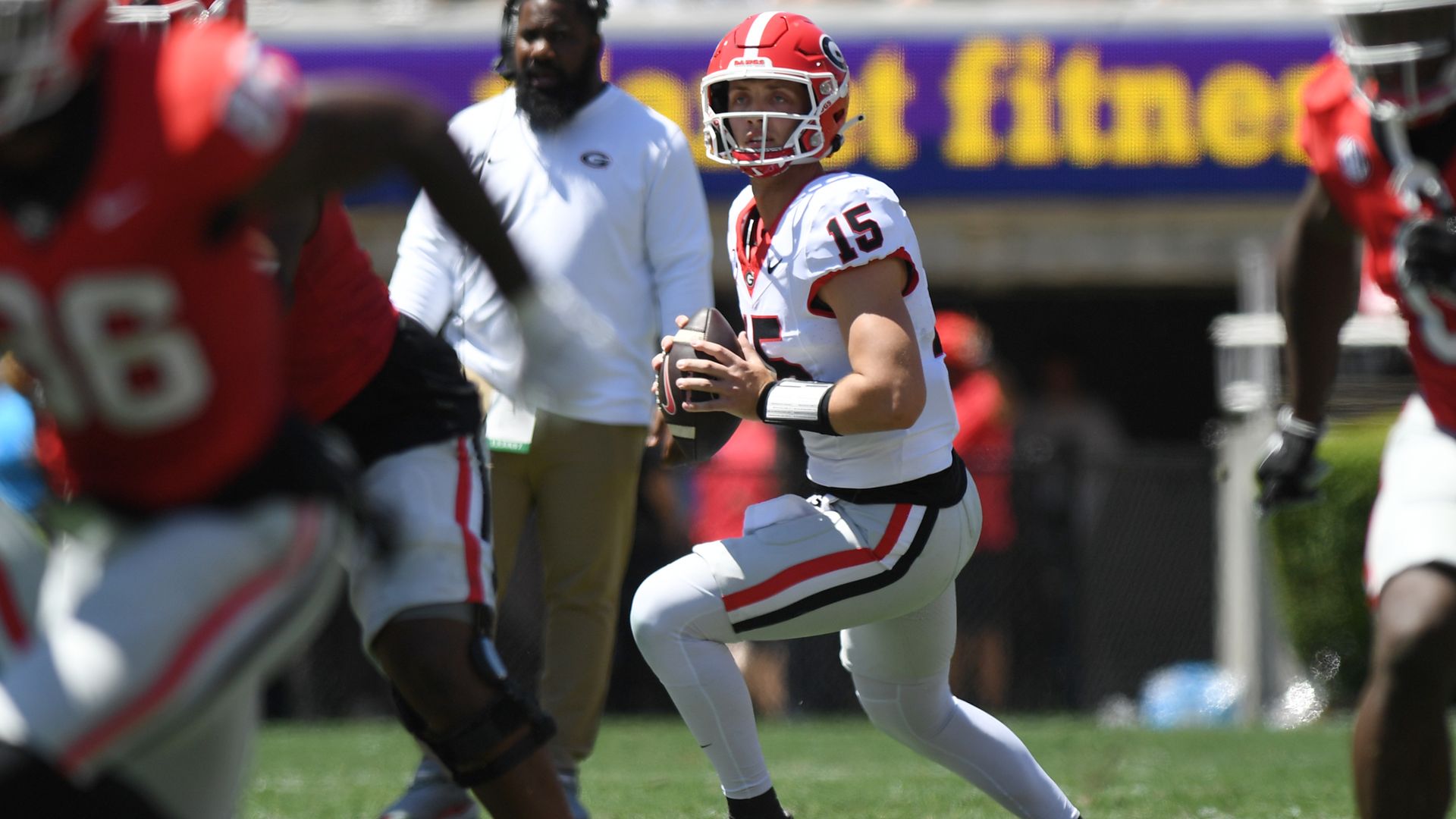 A football quarterback wearing a white uniform and red helmet looks for a teammate to throw a pass during a daytime game