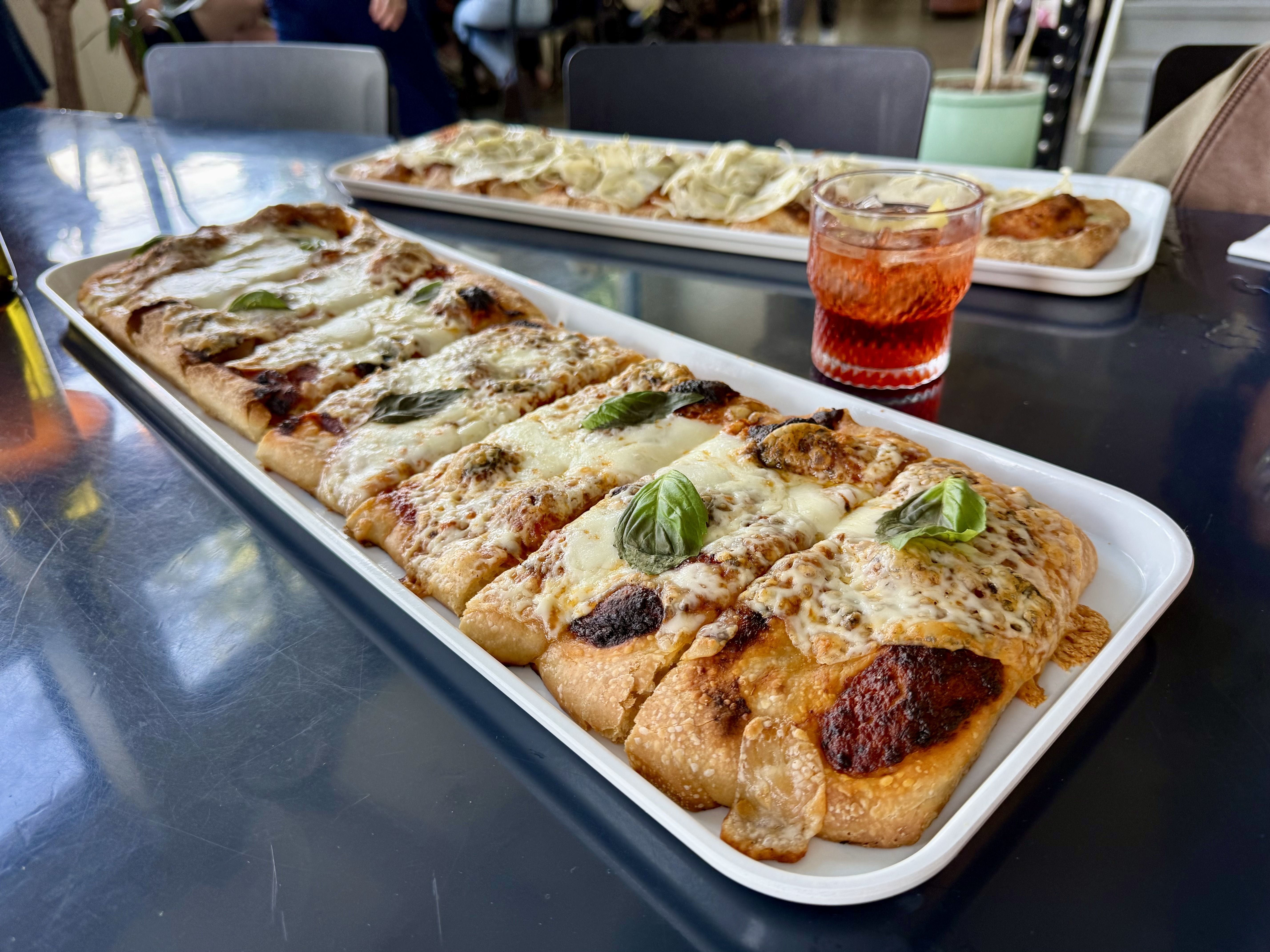 Two rectangular pizzas on white rectangular plates are arranged on a table with a reddish colored Negroni drink in between.