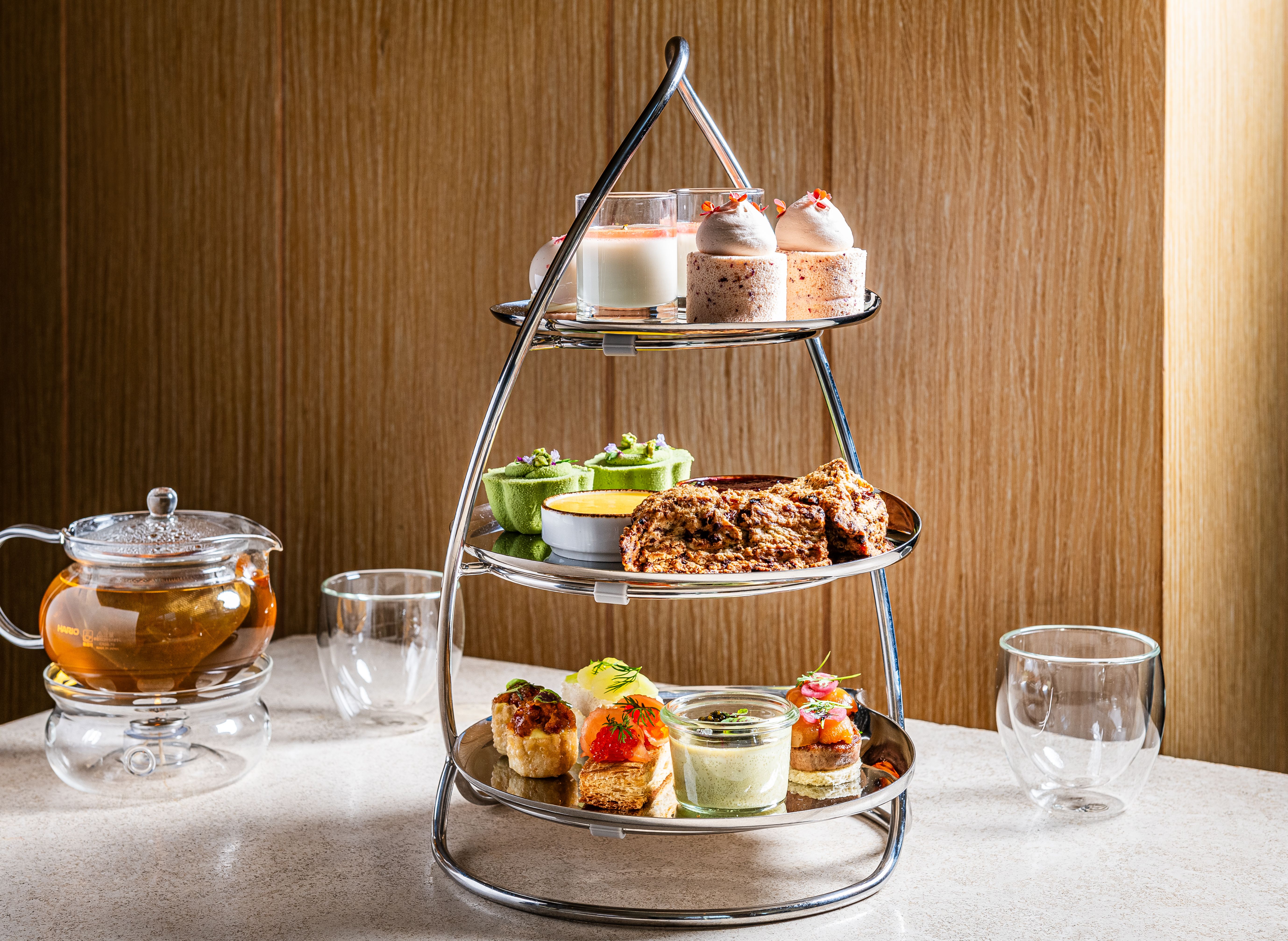 Three-tier metal stand on a white countertop holds assorted treats: top mini cakes with cream, middle green pastries, bottom savory bites. A glass teapot and cups sit nearby against a wood-paneled wall.