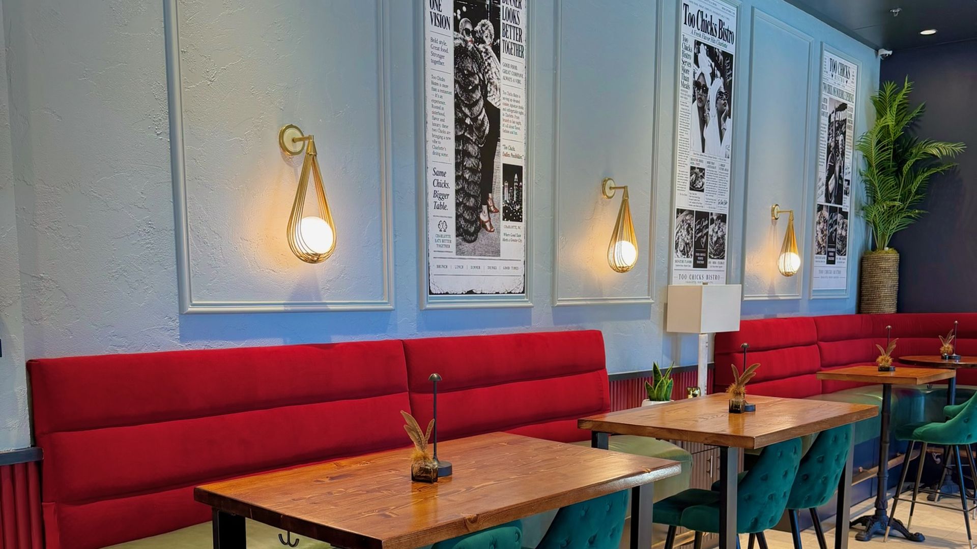 Interior of a cafe with blue paneled walls, framed black-and-white posters titled "Too Chicks Bistro" along the wall, warm gold teardrop lamps, red booth seating, and teal chairs at wooden tables; a potted plant in the corner. Too Chicks restaurant NoDa