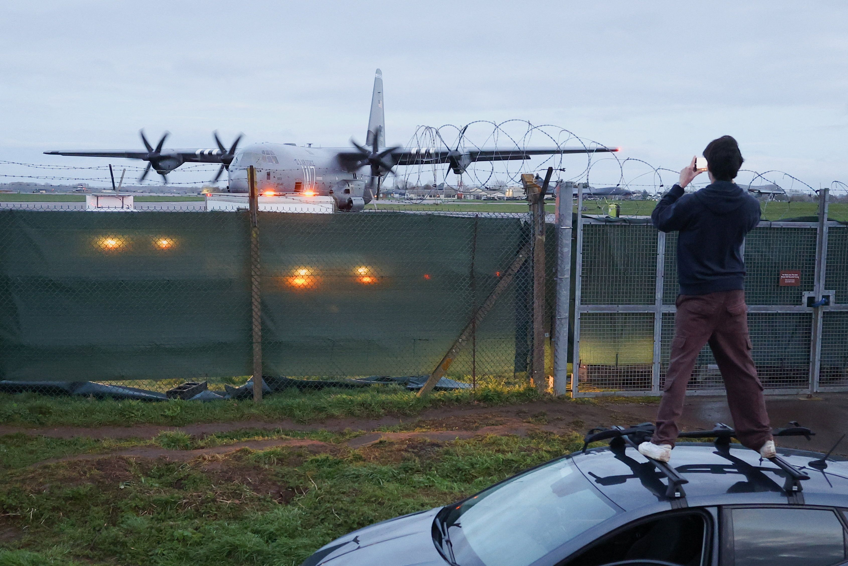 A man stands on the roof of a car to look over temporary screening, placed around a perimeter fence at RAF Fairford airbase, which is used by USAF personnel.