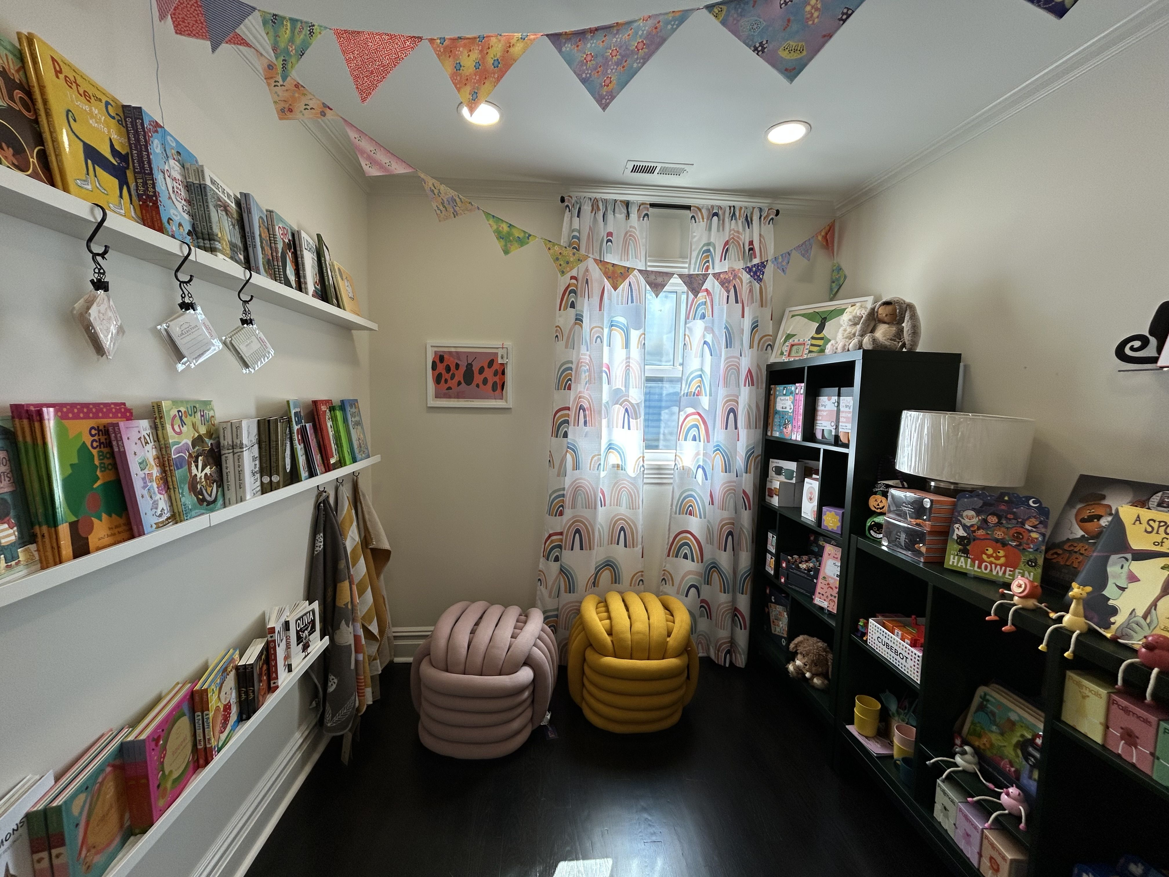 Cozy children's room with colorful triangular bunting, rainbow print curtains, shelves with books and toys, two plush pink and yellow chairs, and dark wood flooring.