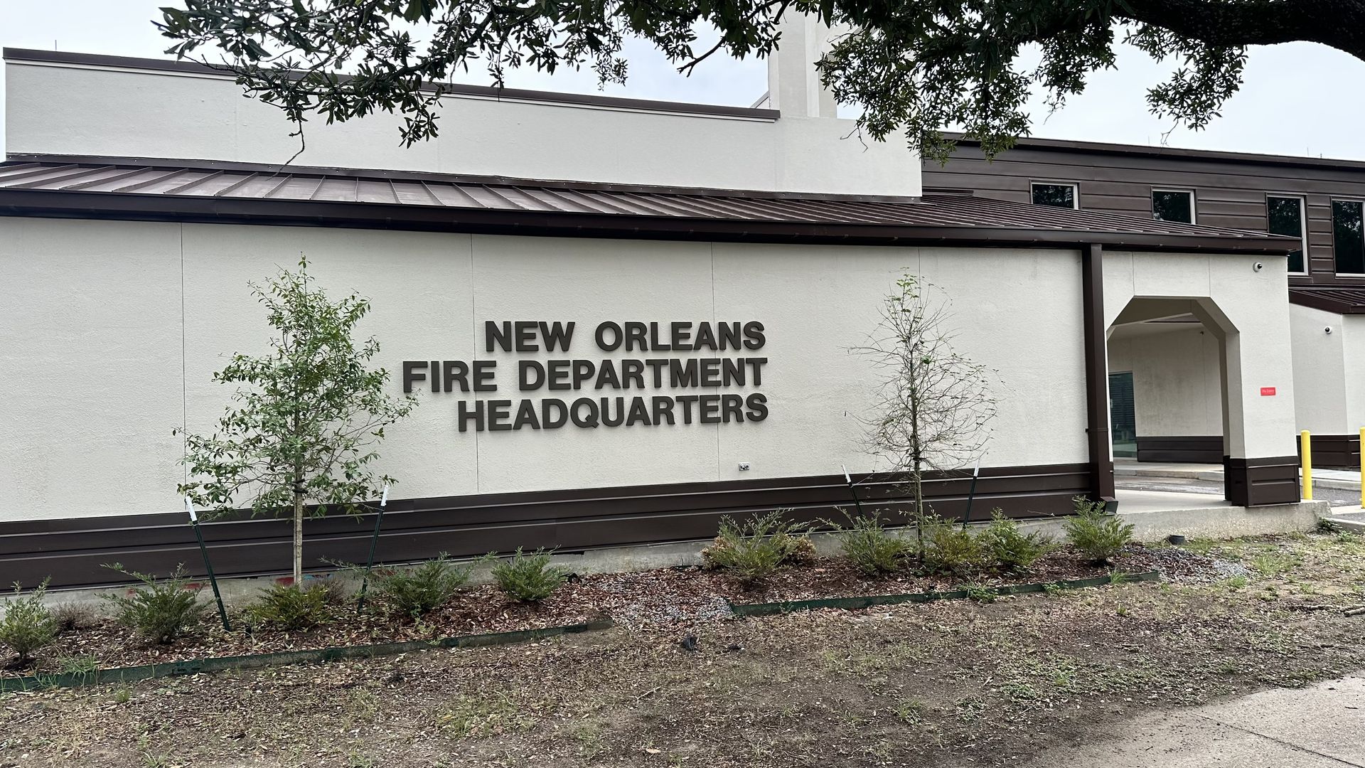 Exterior view of the New Orleans Fire Department Headquarters building: beige walls with brown trim and roof, an arched entry, and a sign reading "New Orleans Fire Department Headquarters" with small trees in front.