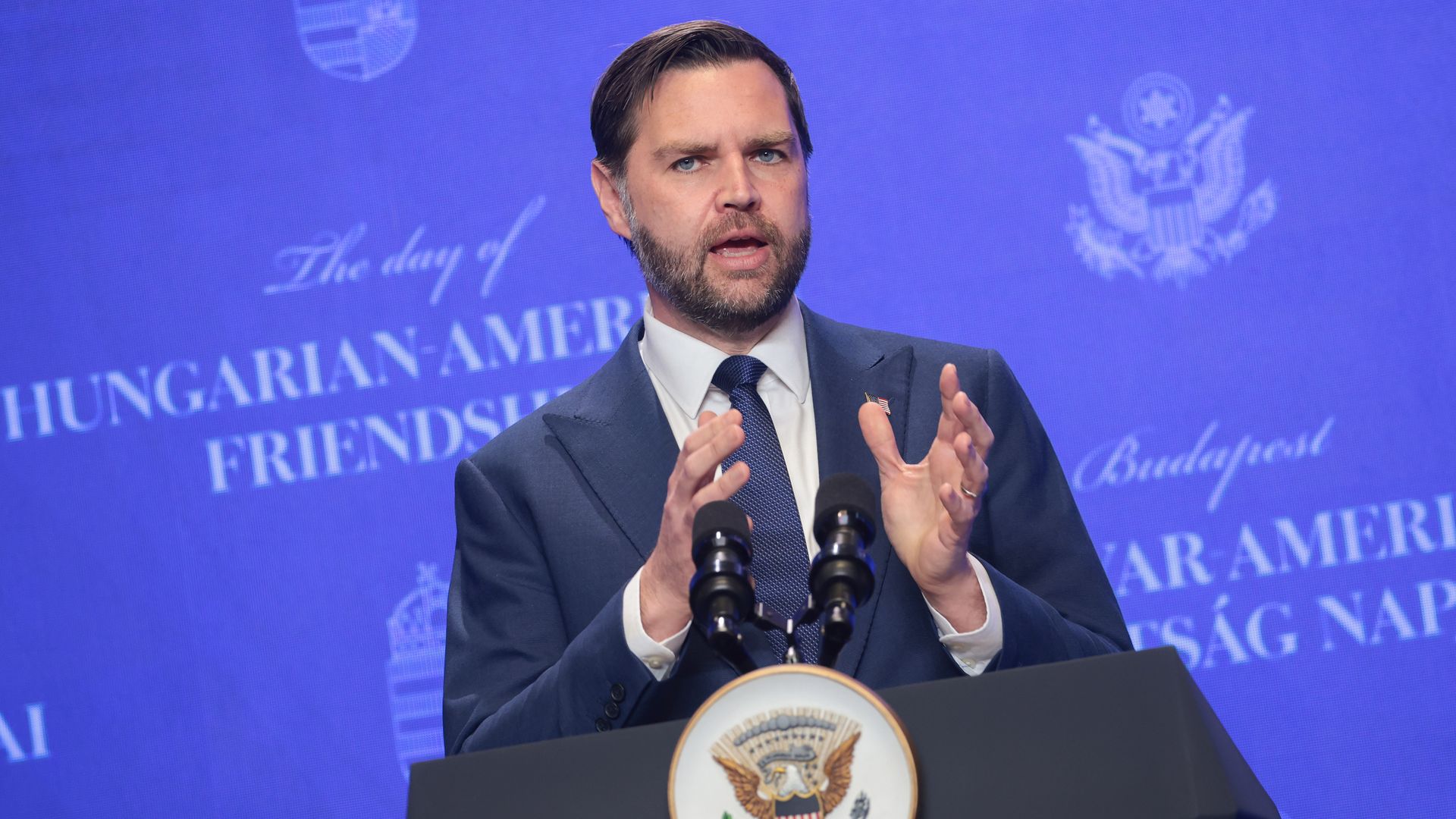 A man in a dark blue suit and tie speaks at a podium with two microphones, gesturing with his hands; a blue backdrop with government seals and text is behind him.
