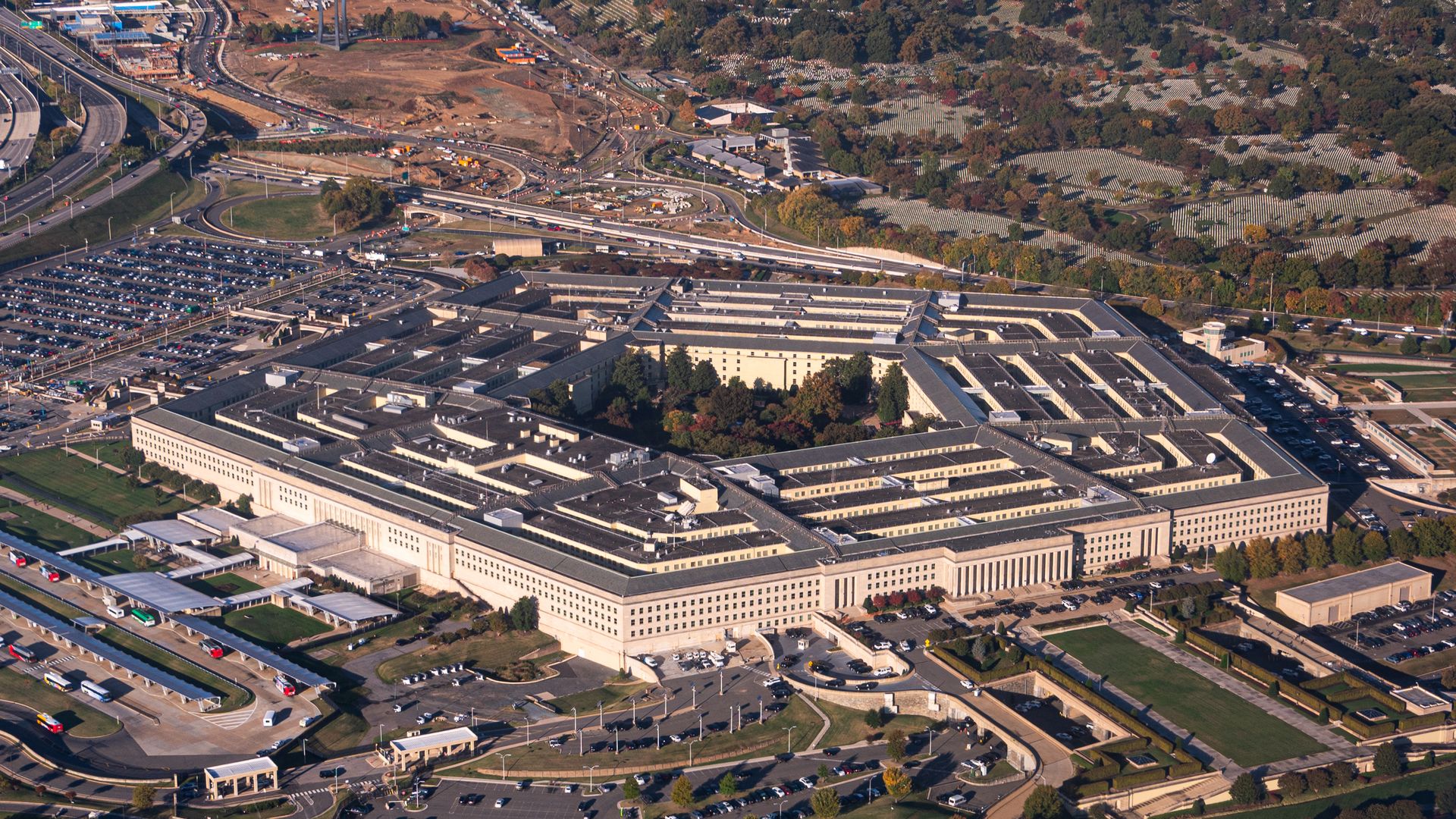Aerial view of the Pentagon building surrounded by parking lots and roads, with trees and urban area in the background under daylight.