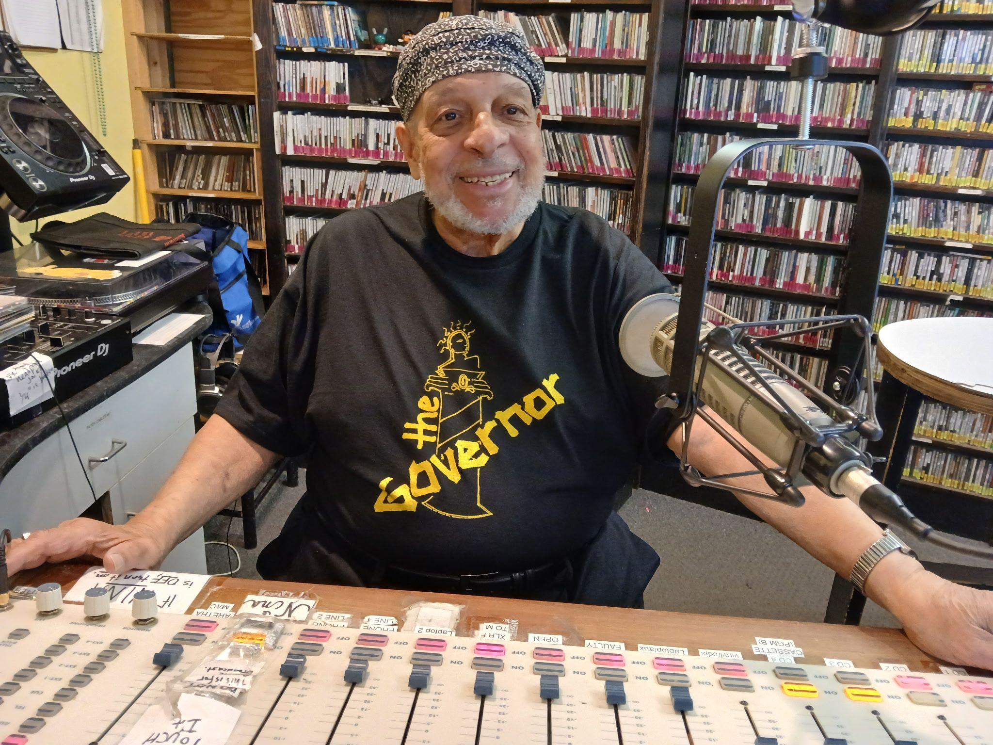 Smiling man wearing a black "the Governor" T-shirt and patterned headscarf sits at a radio mixing console with labeled sliders and a large microphone, surrounded by CDs.