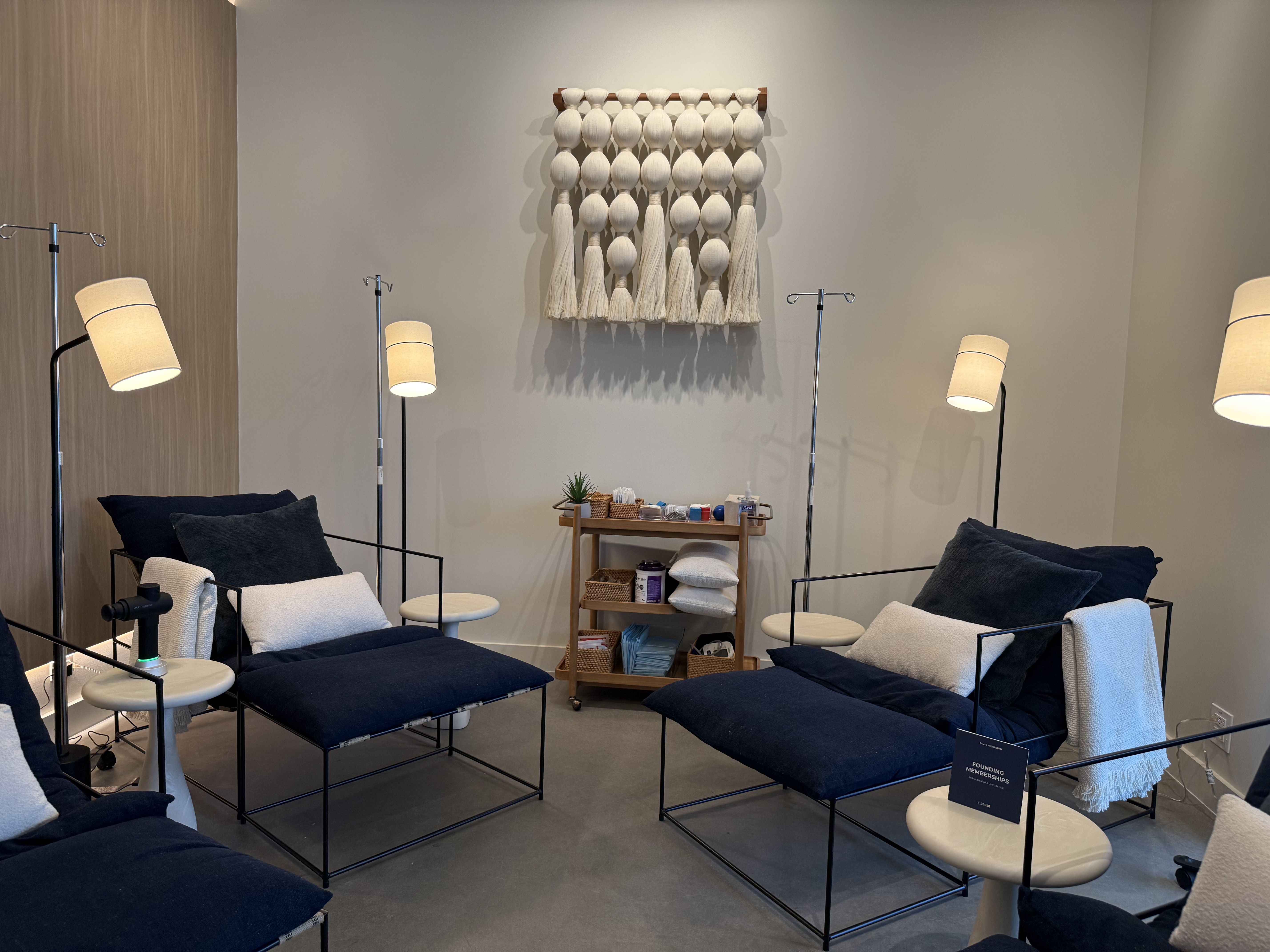 Cozy treatment room with four dark navy cushioned chairs, each with white pillows and blankets, beside small round tables and standing lamps, a wooden cart with supplies, and a woven wall hanging.