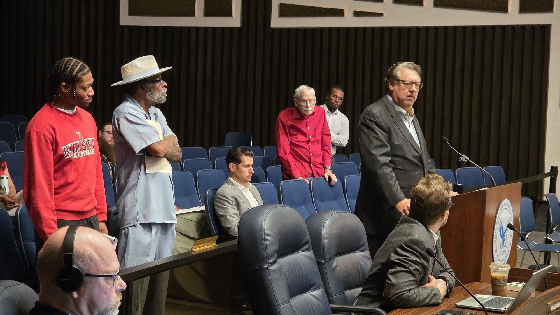 People in a council room: left, a young man with braids wearing a red sweatshirt; next, an older man in light blue attire with a tan hat; center, a man in a red shirt standing; right, a suited man at a podium.