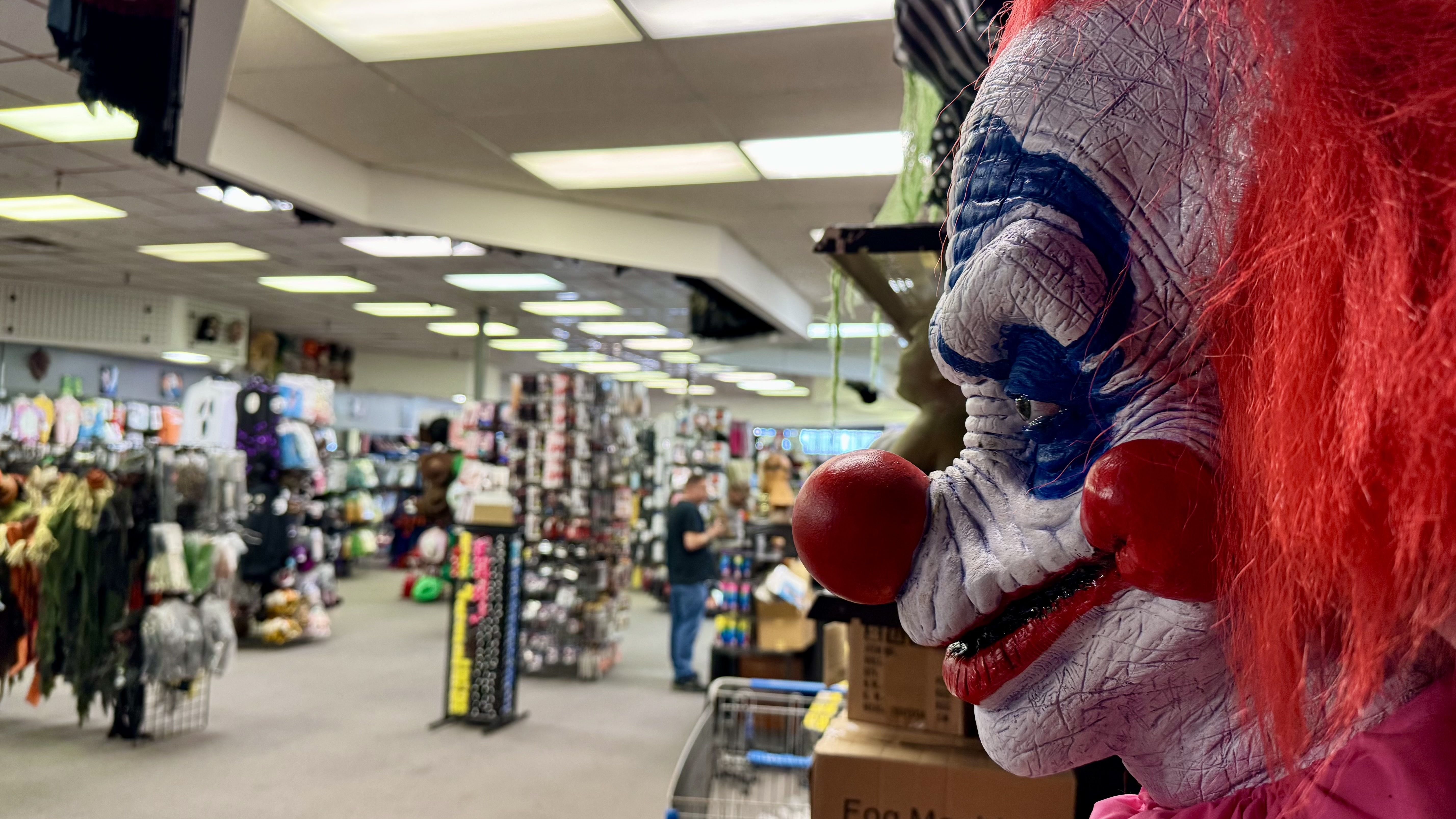 A life size clown figurine is pictured in the foreground with the halloween store offerings in the background