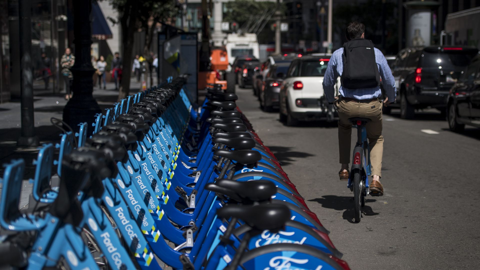 Photo of a person riding an electric bike past a row of blue electric bikes parked on the side of the road