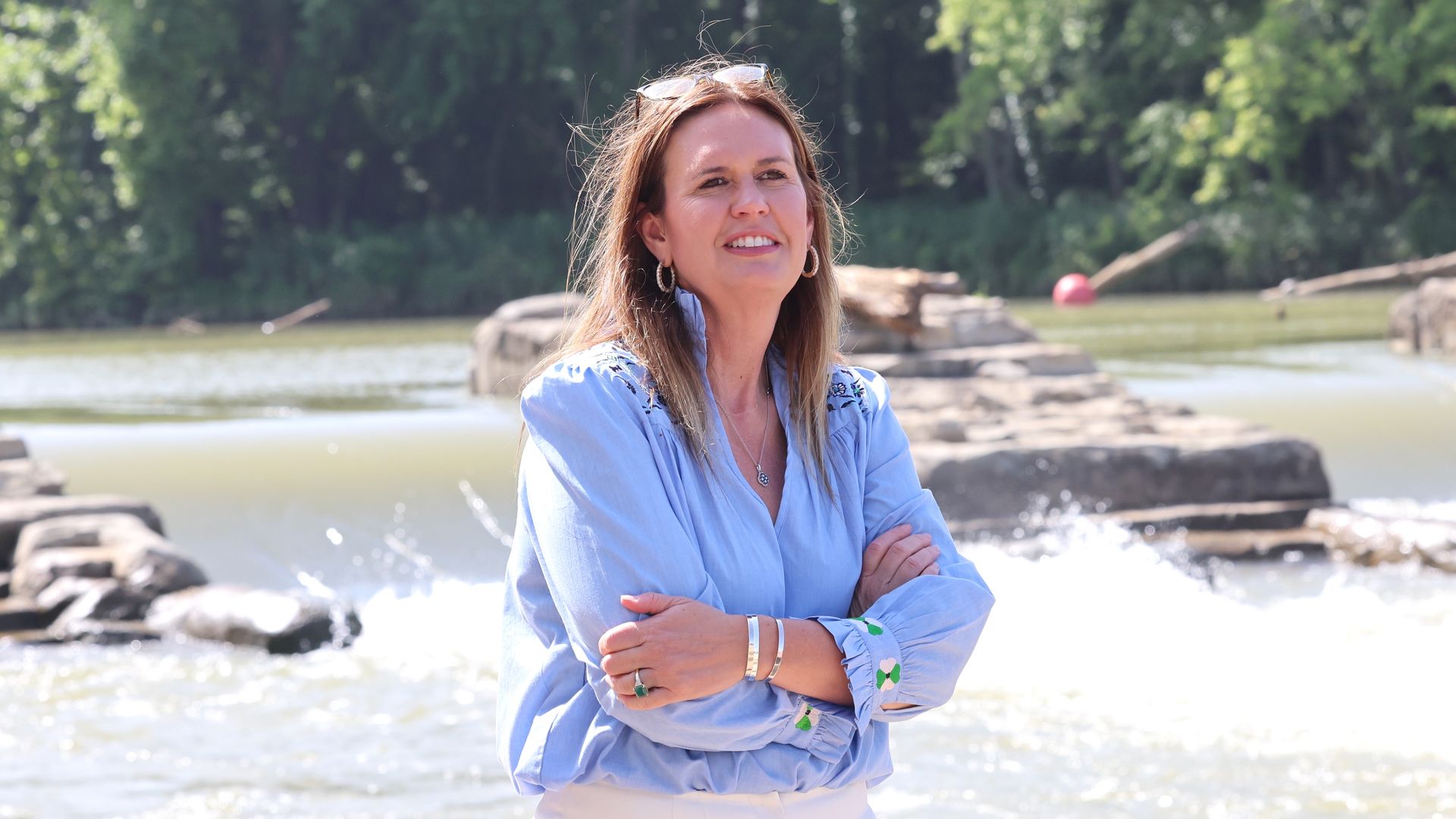 Relaxed woman stands on a rocky riverside with trees in the background, wearing a light blue blouse, white pants, hoop earrings, sunglasses on her head, and a smile with arms crossed.