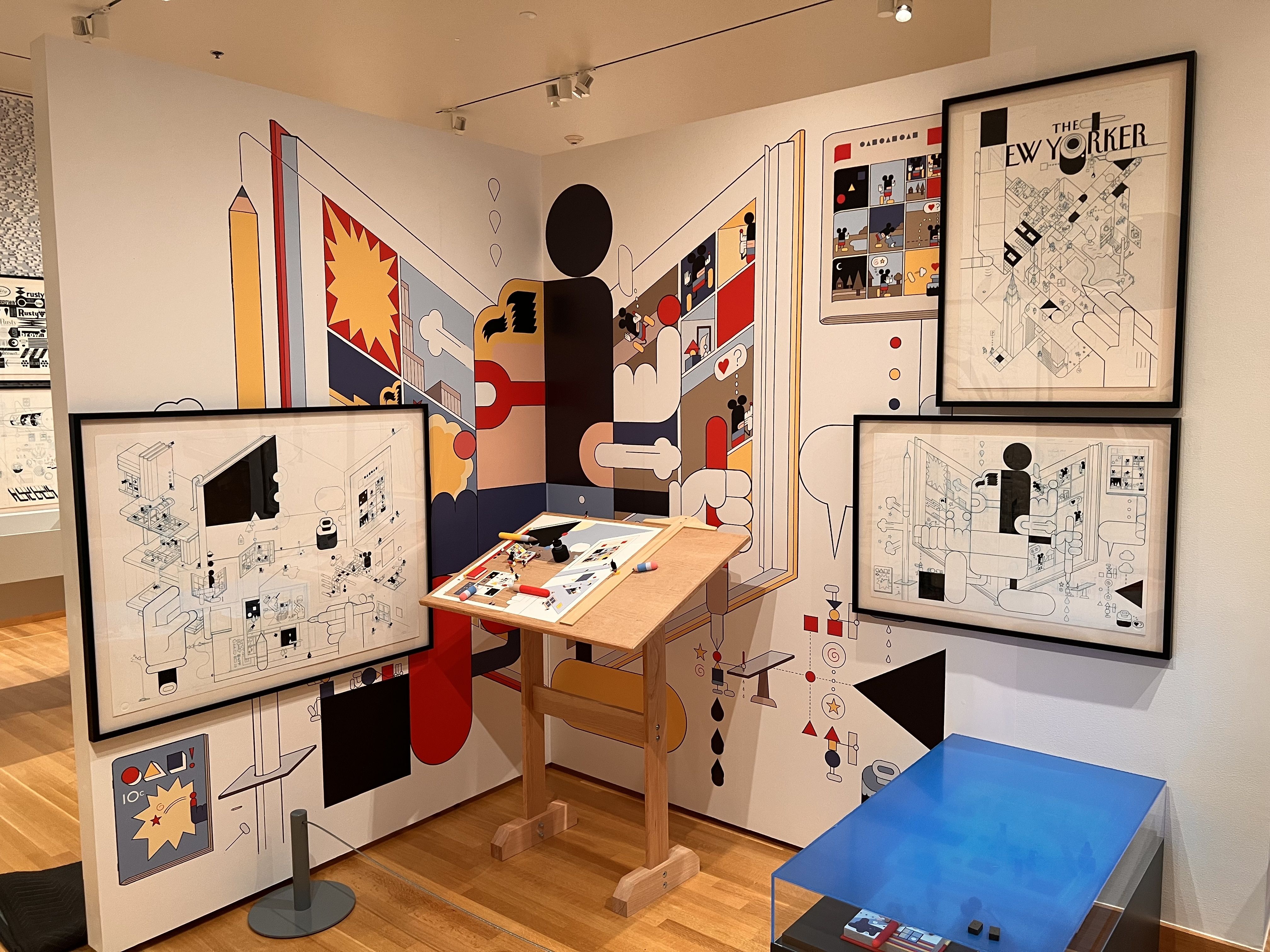 Gallery corner with oversized graphic panels of abstract geometric art in red, yellow and blue; a framed "The New Yorker" print; a wooden drafting table with markers; and blue benches nearby.