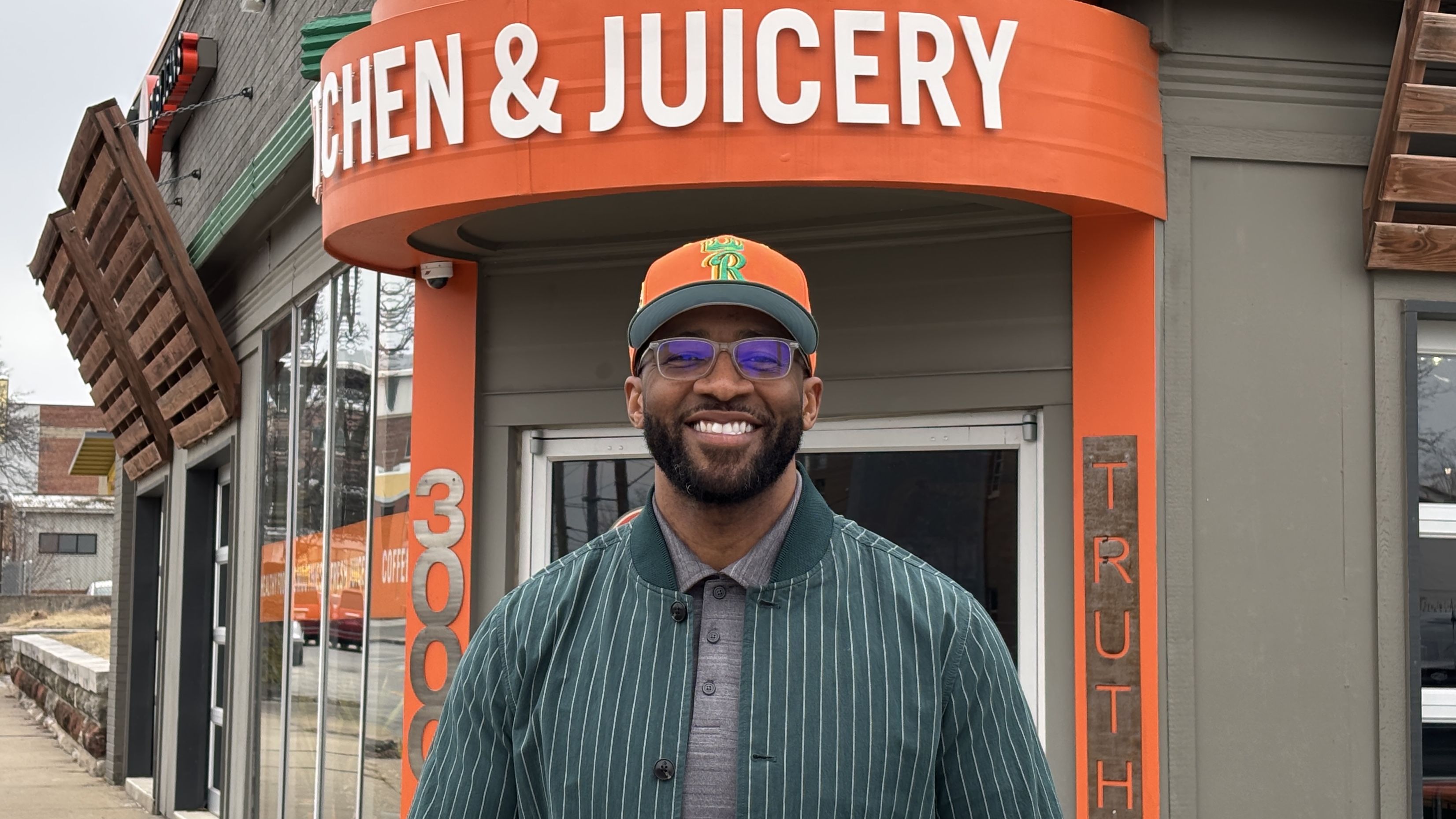 Chris Goode wearing an orange and green cap, glasses, green striped jacket, standing outside his Ruby Jean's Juicery storefront on Troost Avenue with orange signage reading "KITCHEN & JUICERY" and "TRUTH".
