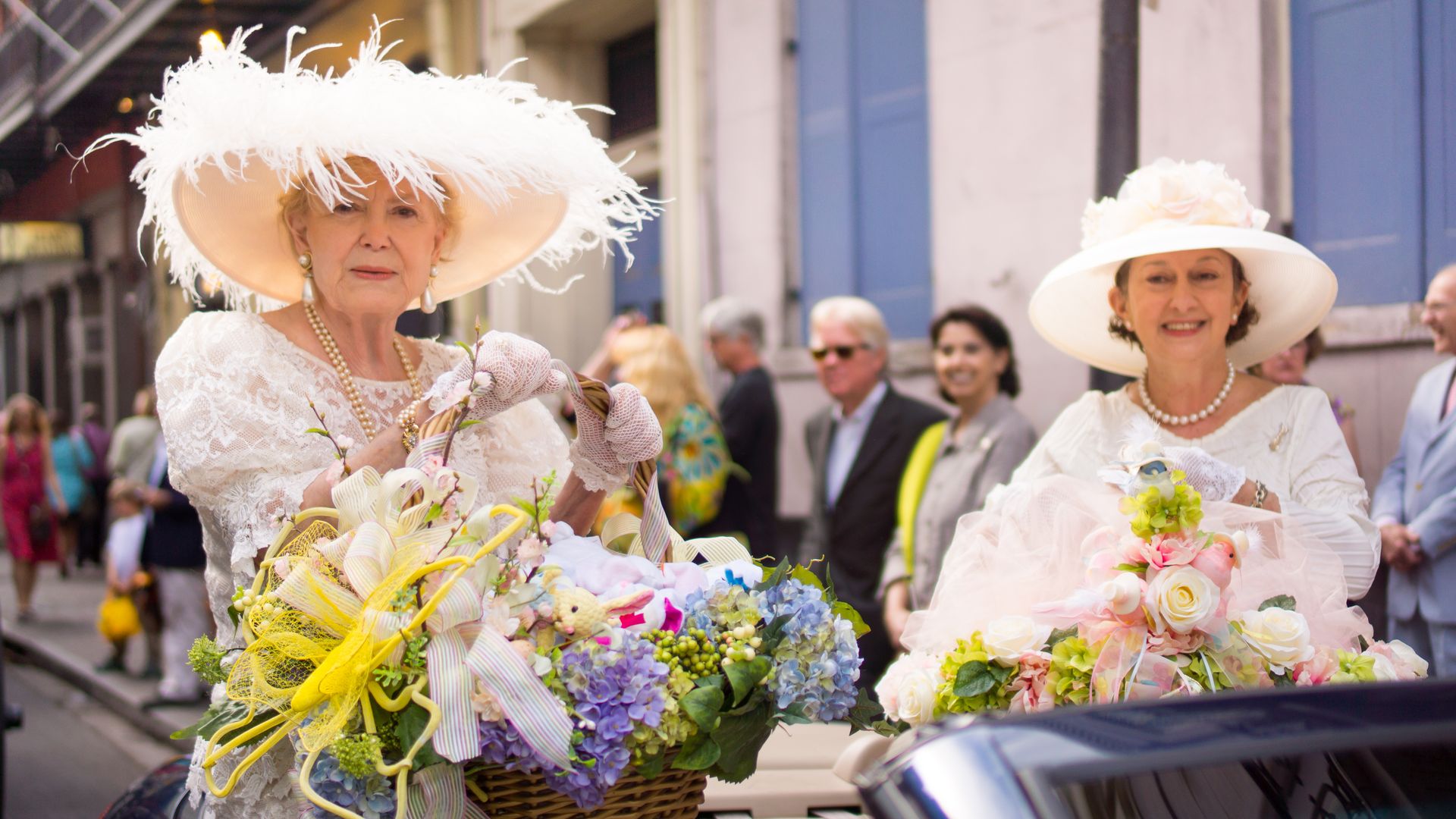 Image shows two women with white Easter bonnets and baskets of flowers sitting on a convertible car.