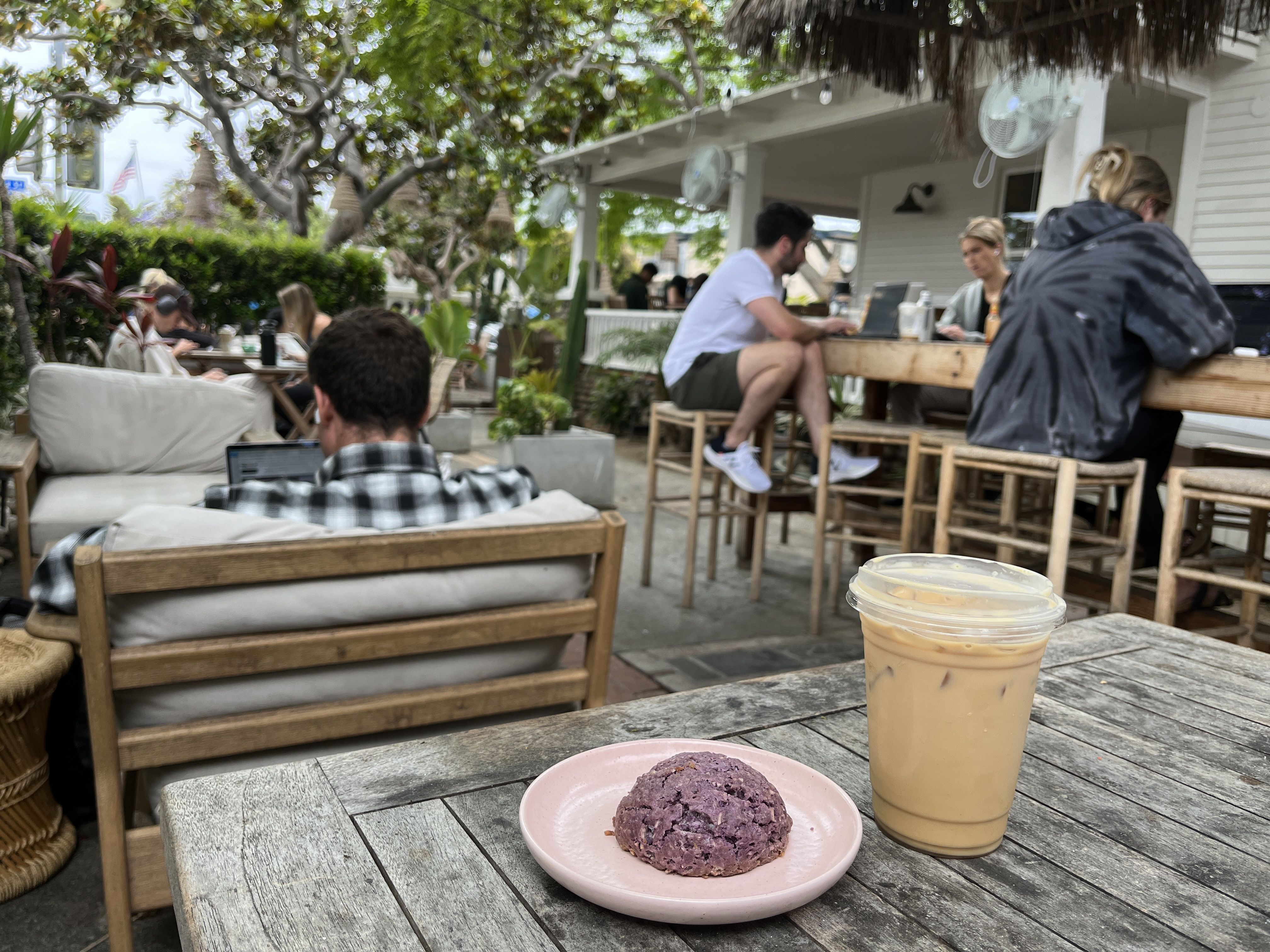 An iced coffee and a purple coconut scone sit on a table in the outdoor garden of a coffee shop where people are working at nearby tables. 