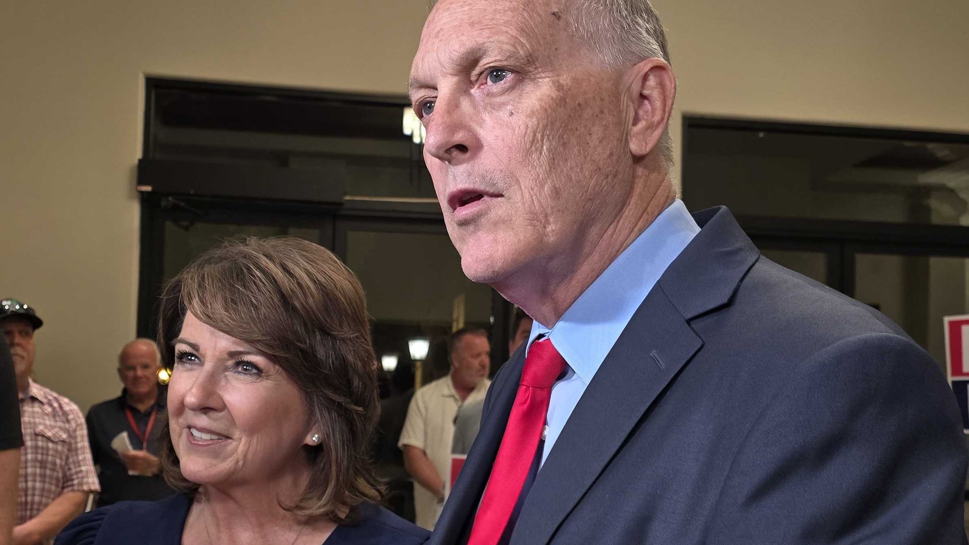 Sine Kerr and Andy Biggs at a political event: Biggs in a blue suit and red tie, and a smiling Kerr in navy, surrounded by microphones; signs in the background read "BIGGS" and "KERR".