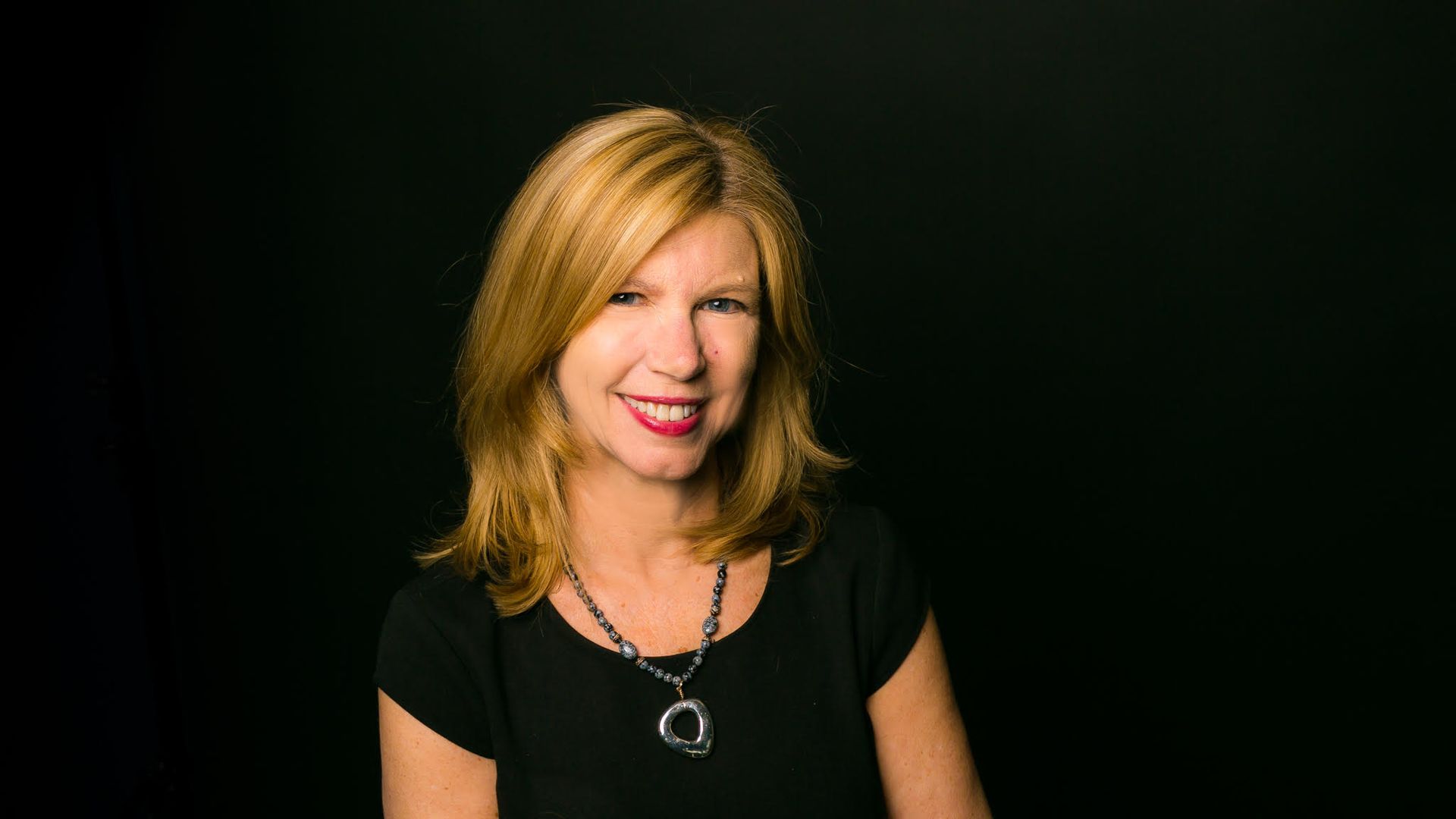 Smiling blonde woman with shoulder-length hair wearing a black top and a necklace with a large silver pendant, standing against a dark background.