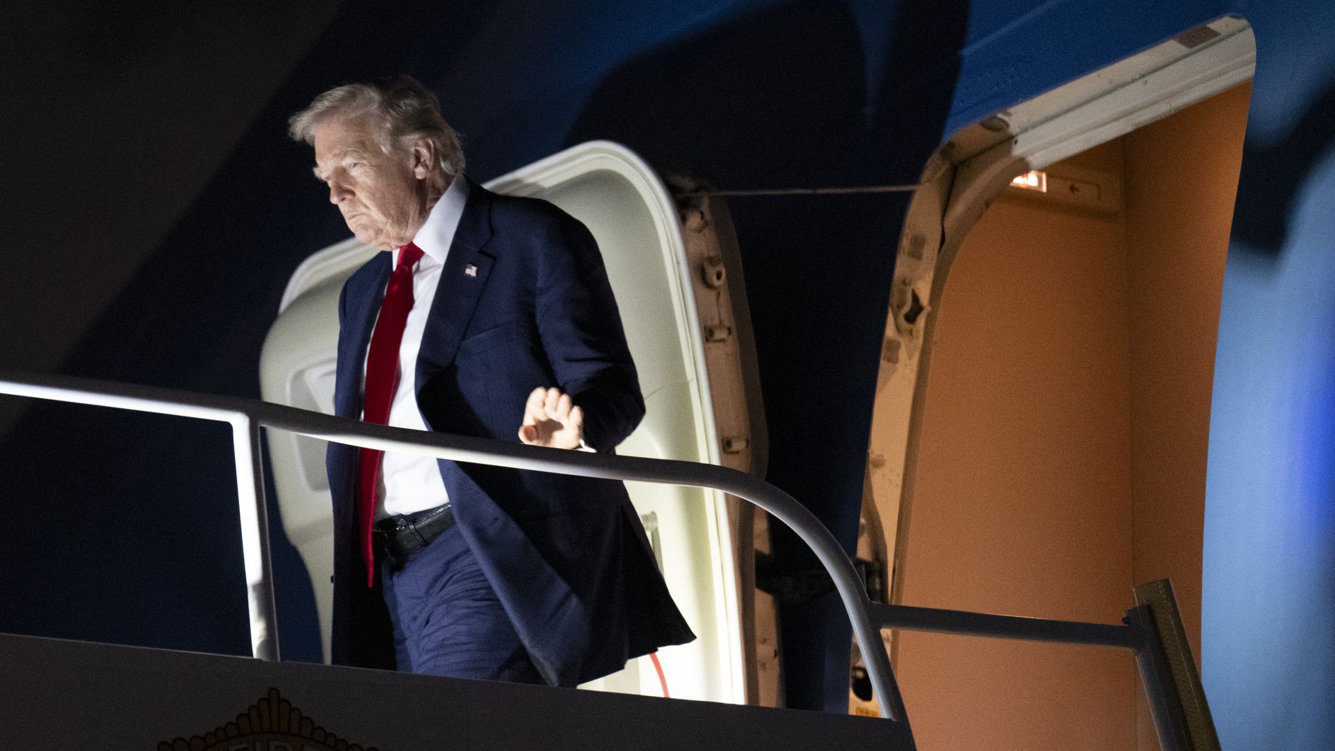 President Trump, illuminated by footlights against a dark background, exits an airplane. 