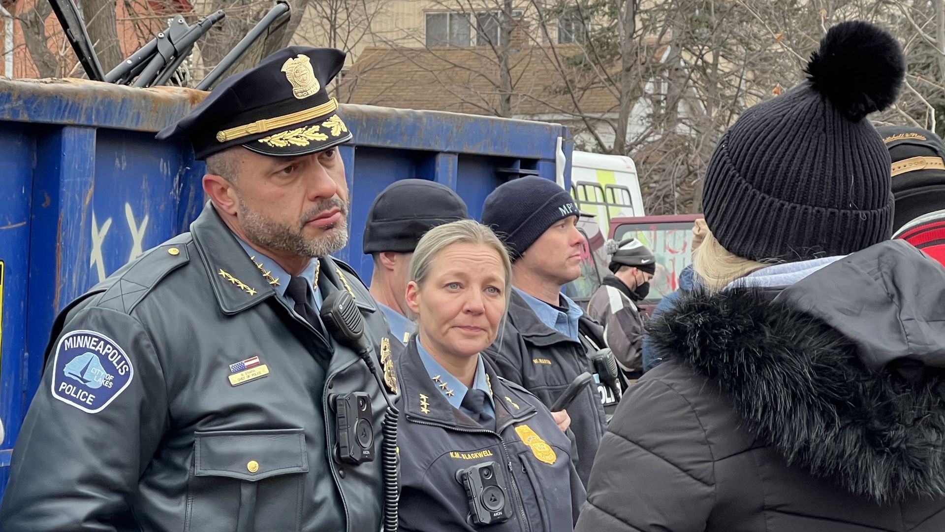 Minneapolis police officers in uniform stand beside a blue dumpster, engaging with a group of people dressed in winter jackets and hats in a cold outdoor setting.