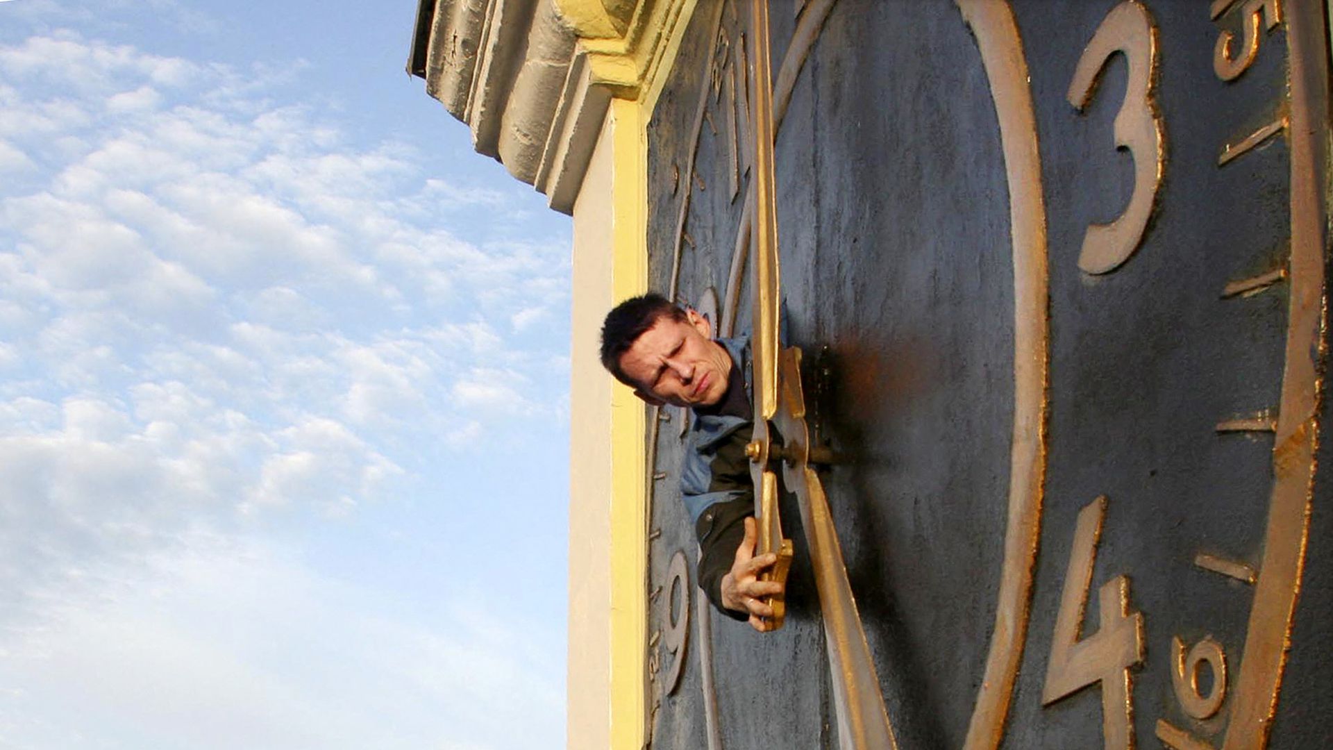 A person's head pokes out of the face of a huge clock in a tower against a slightly cloudy sky. 