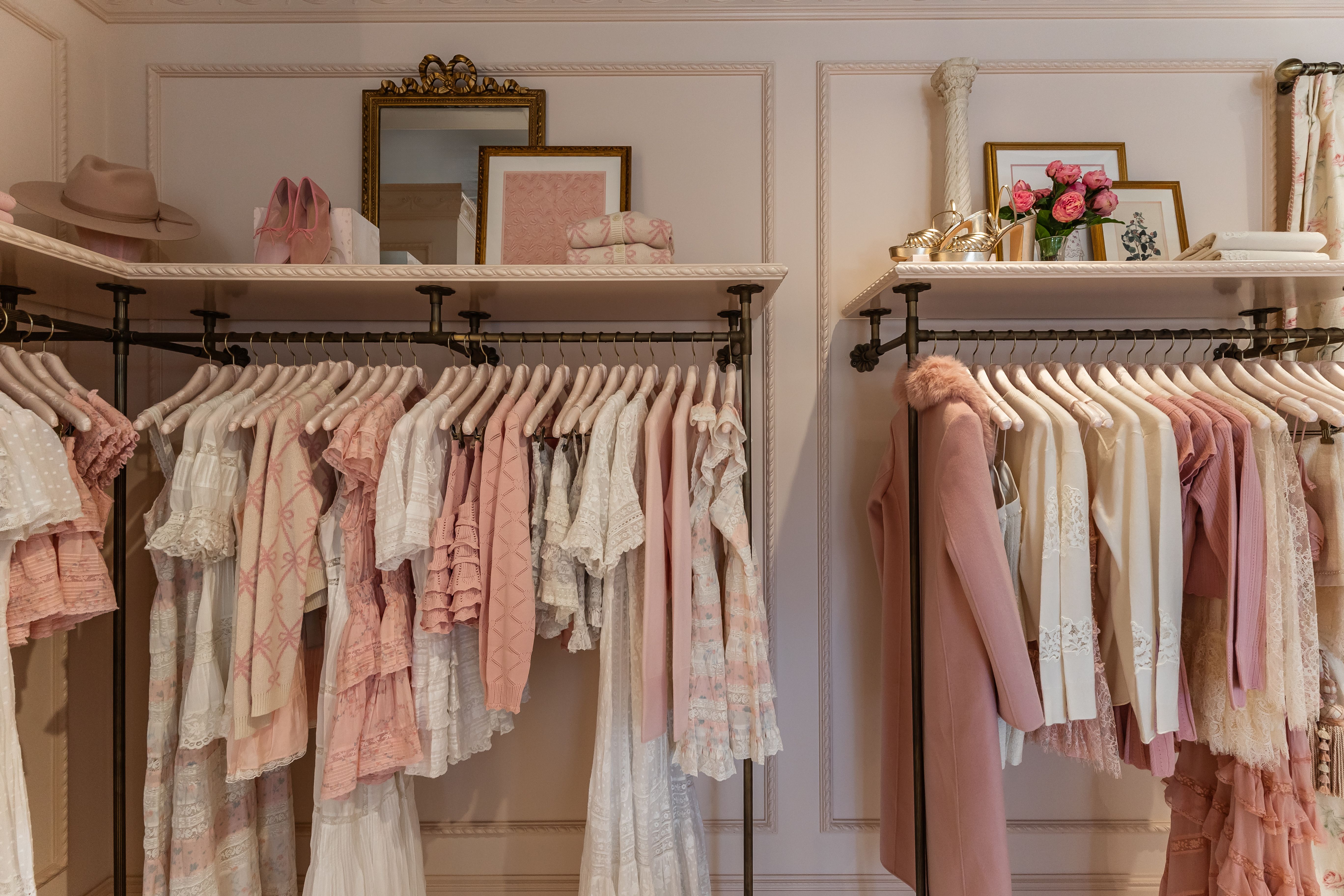 Clothing racks with pastel dresses, coats, and tops in pink and white tones, a beige hat, pink shoes, gold sandals, framed art, and a vase of pink roses on shelves in a softly lit room.