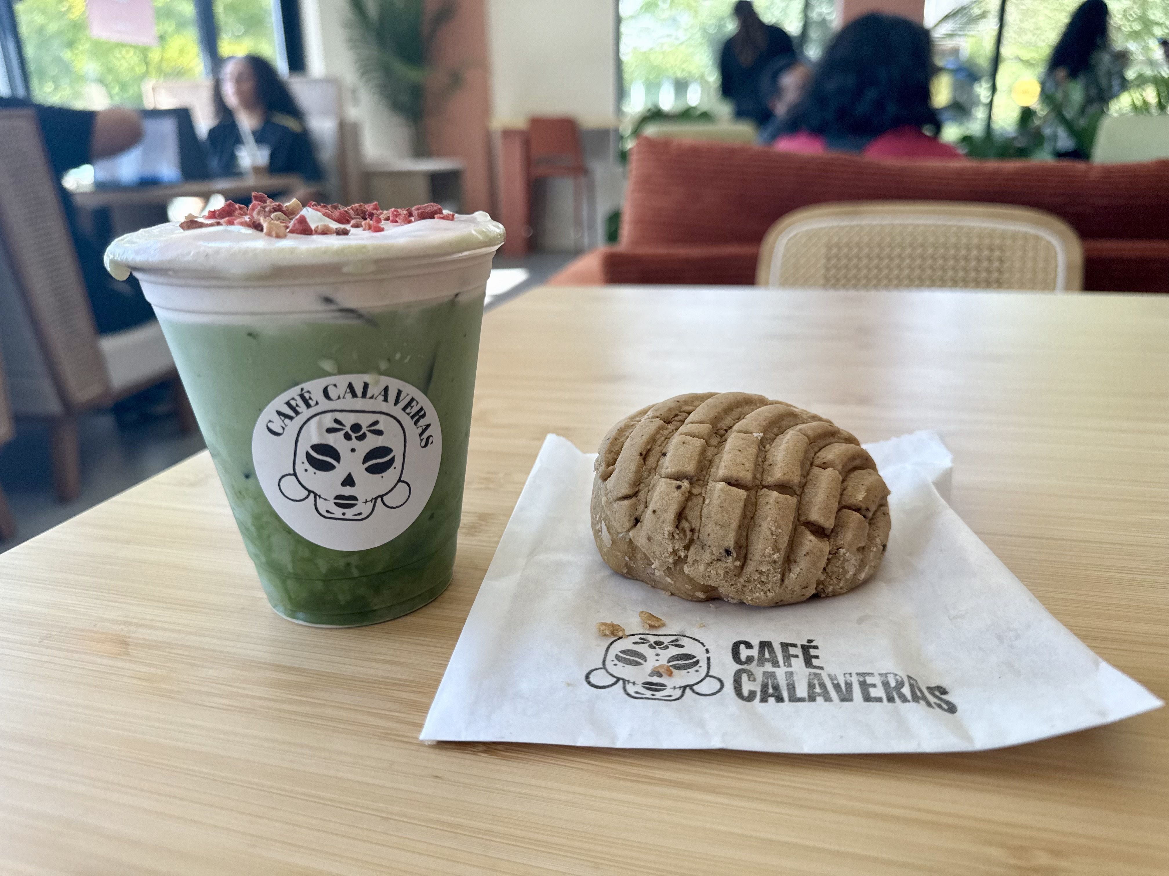 Iced green matcha drink with cold foam and red dried strawberries on top in a clear cup labeled "Café Calaveras" next to a brown concha pastry on a white "Café Calaveras" napkin, on a wooden table in a cafe.