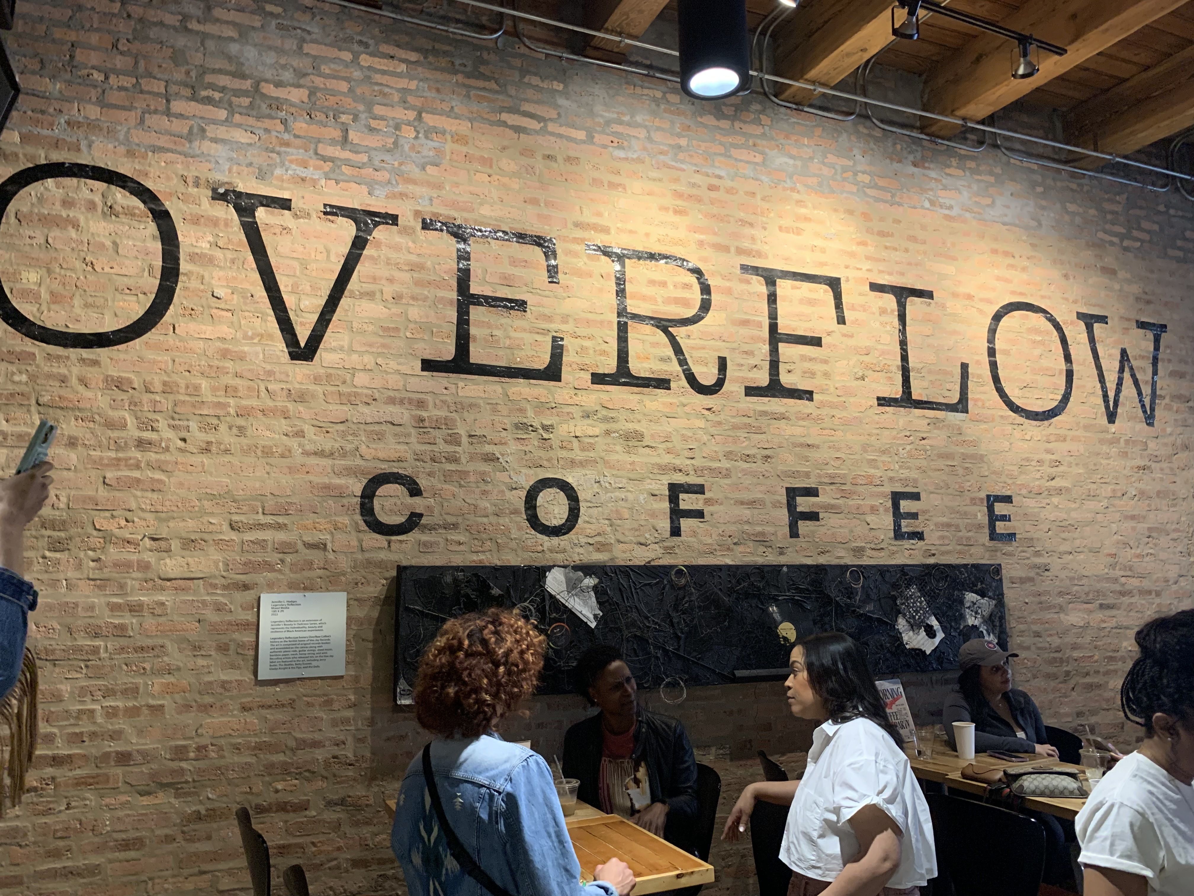 Interior of Overflow Coffee shop with brick wall featuring large black letters spelling "OVERFLOW COFFEE." Several people are seated and chatting at wooden tables under ceiling lights.