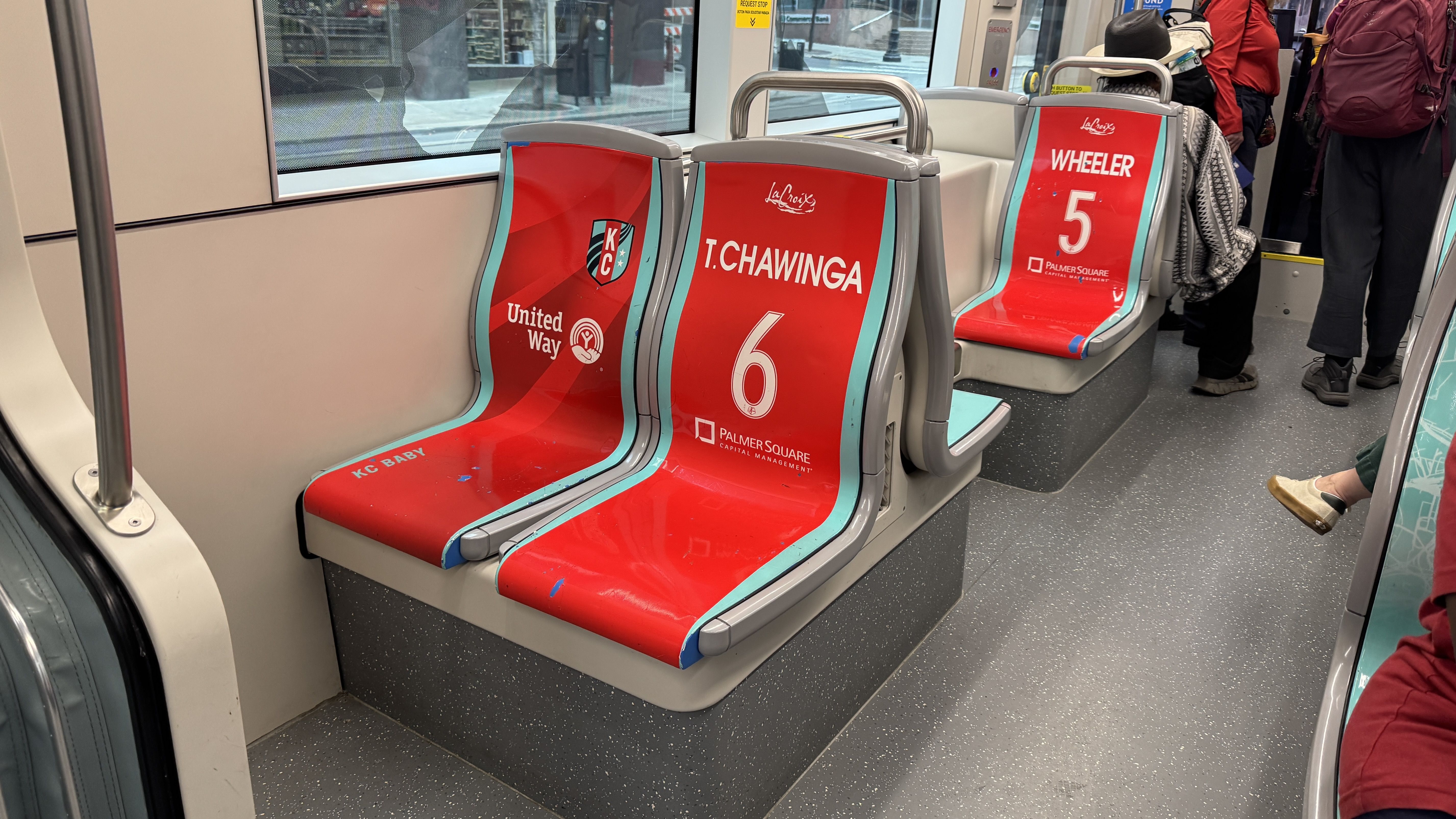 Inside the KC Streetcar with red seats outlined in light blue. Seat 6 reads "T. CHAWINGA," seat 5 reads "WHEELER." Passengers stand by the door near a window.