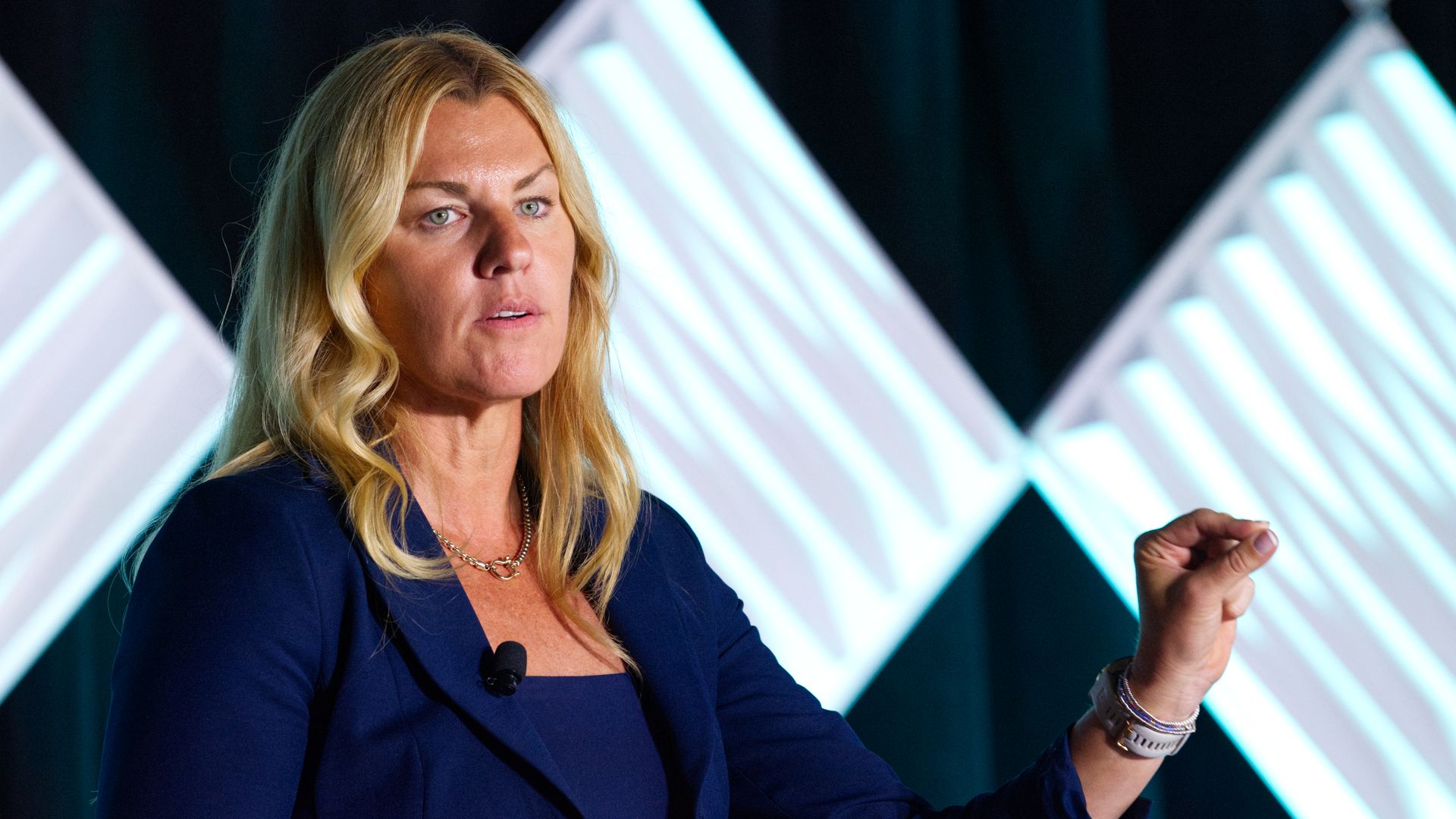 Blonde woman in a navy blue blazer speaking with a microphone clipped to her jacket, gesturing with her right hand, against a dark background with illuminated geometric panels.
