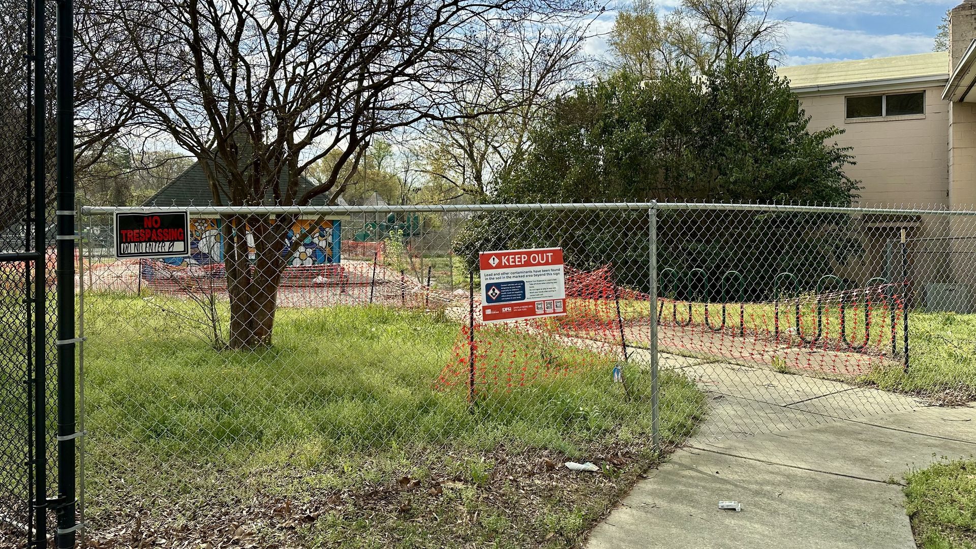 Chain-link fence surrounds a vacant lot with a large bare tree, orange safety netting, and a beige building. Red "KEEP OUT" and "NO TRESPASSING" signs stand near a curved sidewalk.