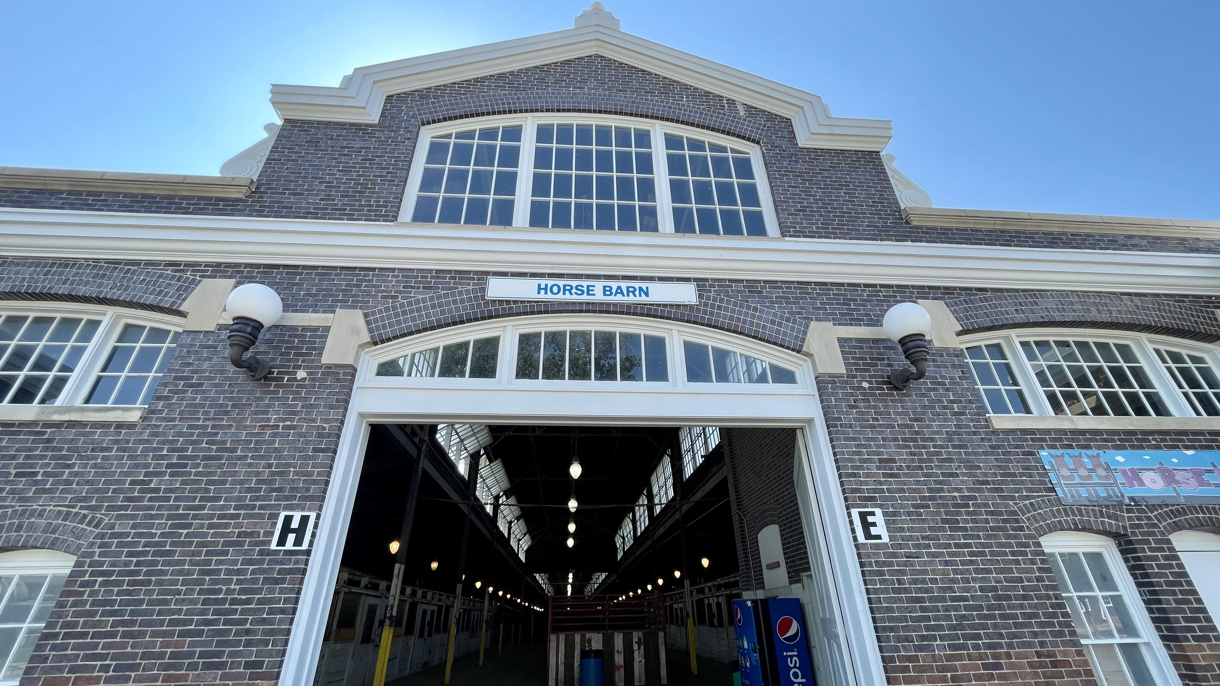 Brick building entrance labeled "HORSE BARN" with large windows and lamps, showing the inside of a barn with rows of stalls and a Pepsi vending machine on the right.
