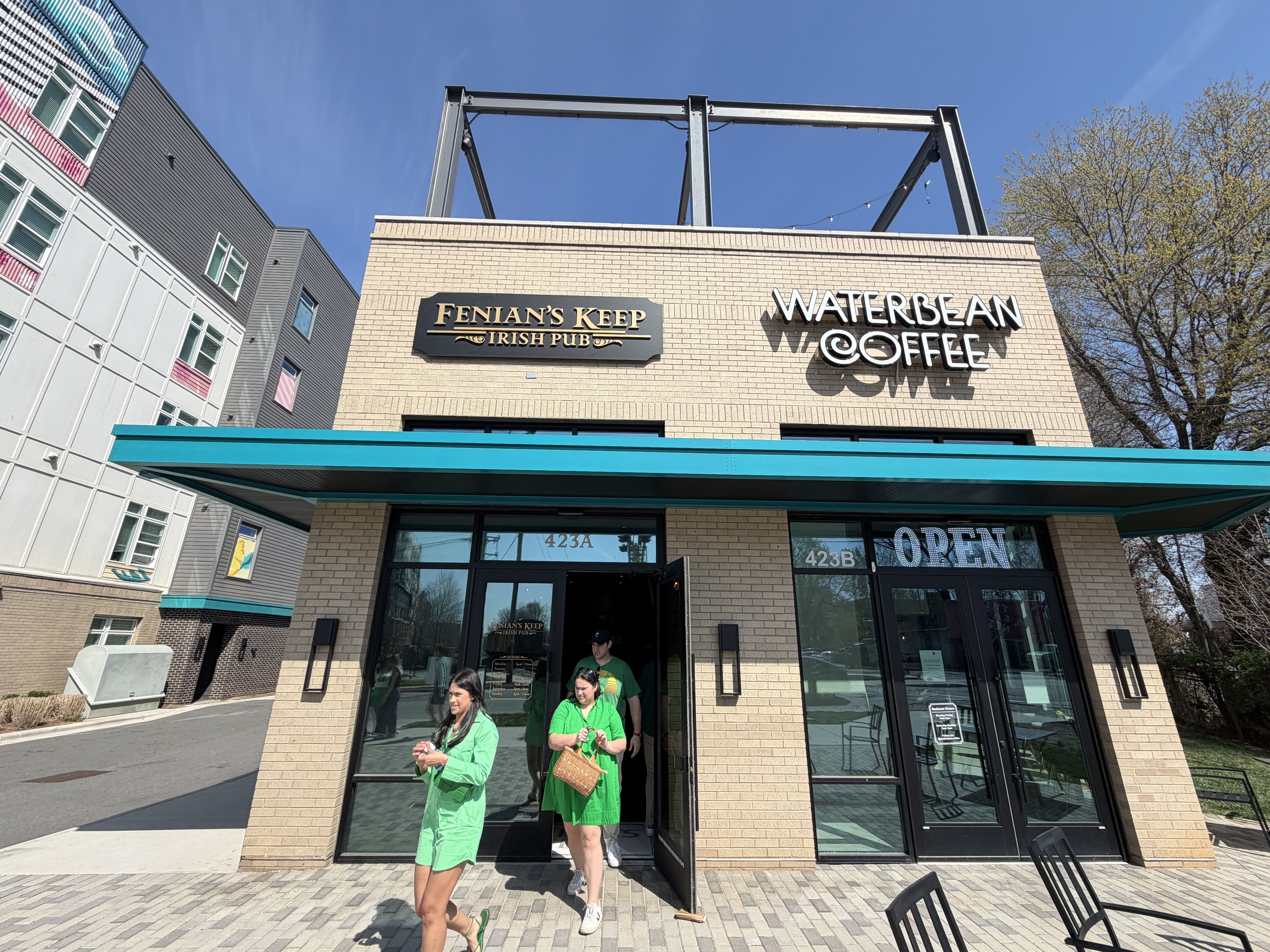 Brick building with a turquoise awning housing Fenian's Keep Irish Pub and Waterbean Coffee. Two women in green exit the entrance; a large OPEN sign and glass windows reflect a sunny day.