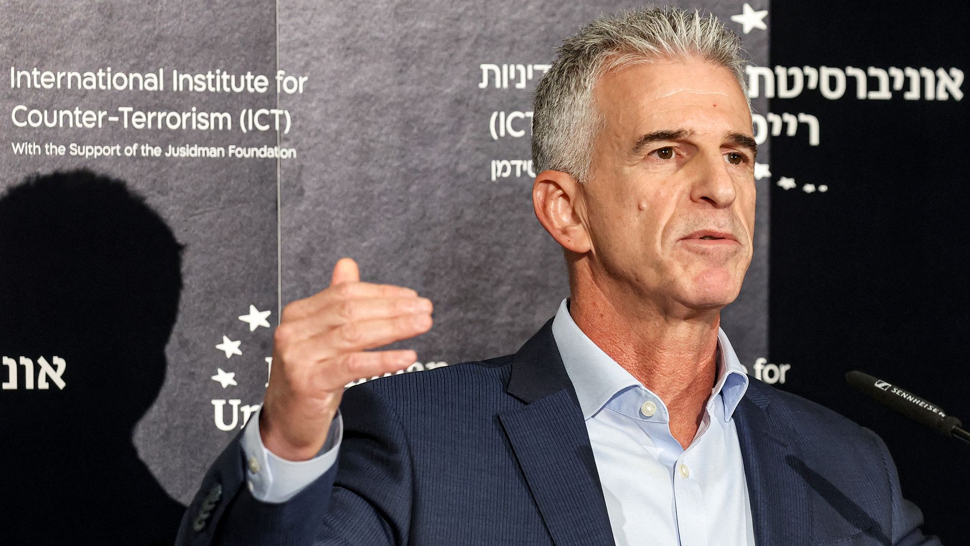 Man in a blue suit and light blue shirt speaking and gesturing with his hand against a backdrop with the International Institute for Counter-Terrorism (ICT) logo in English and Hebrew.