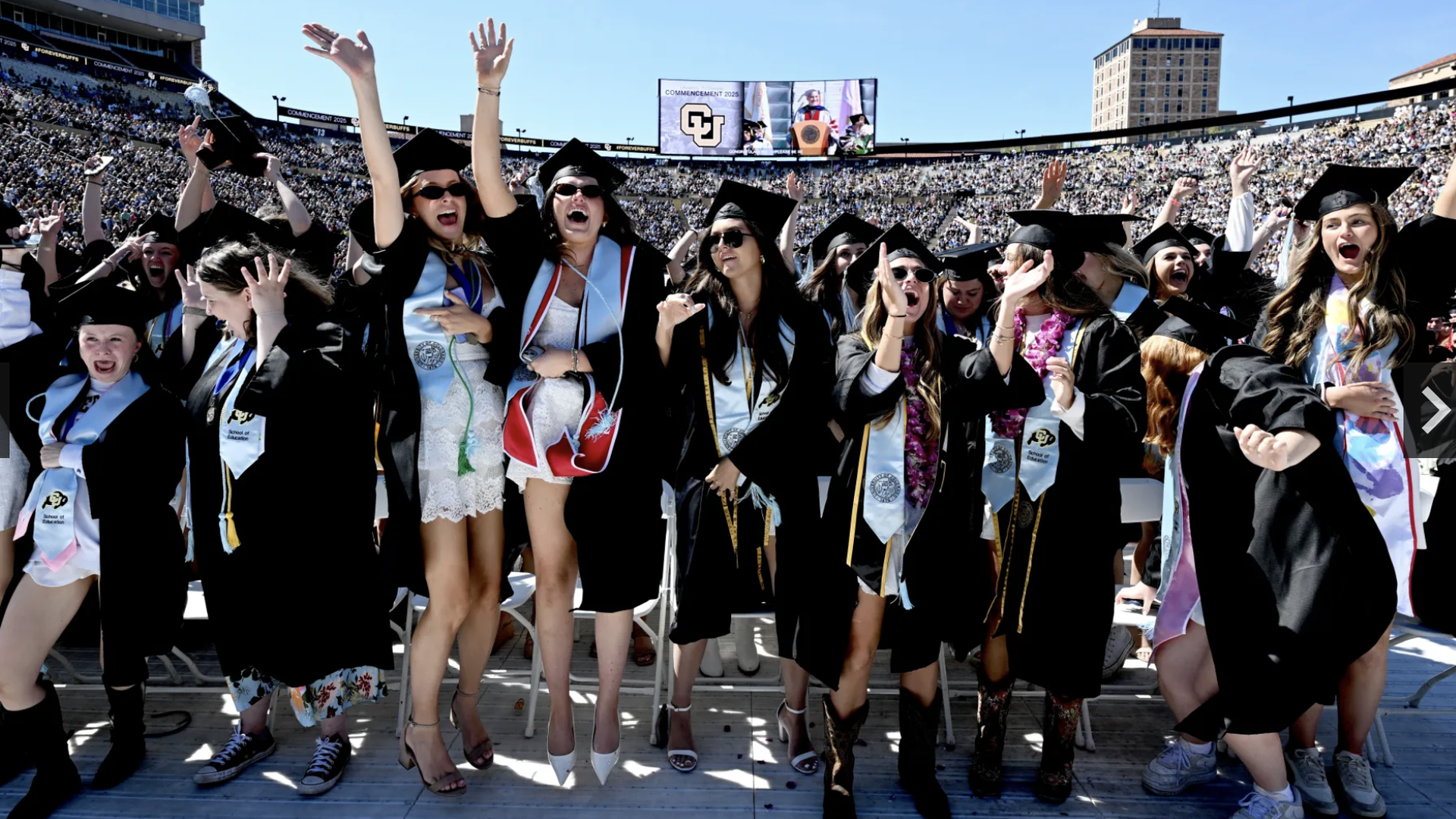 Graduates in black gowns and mortarboard caps cheer with raised arms in a packed stadium under a blue sky; many wear light-blue sashes and leis as a large ceremony screen looms in the background.