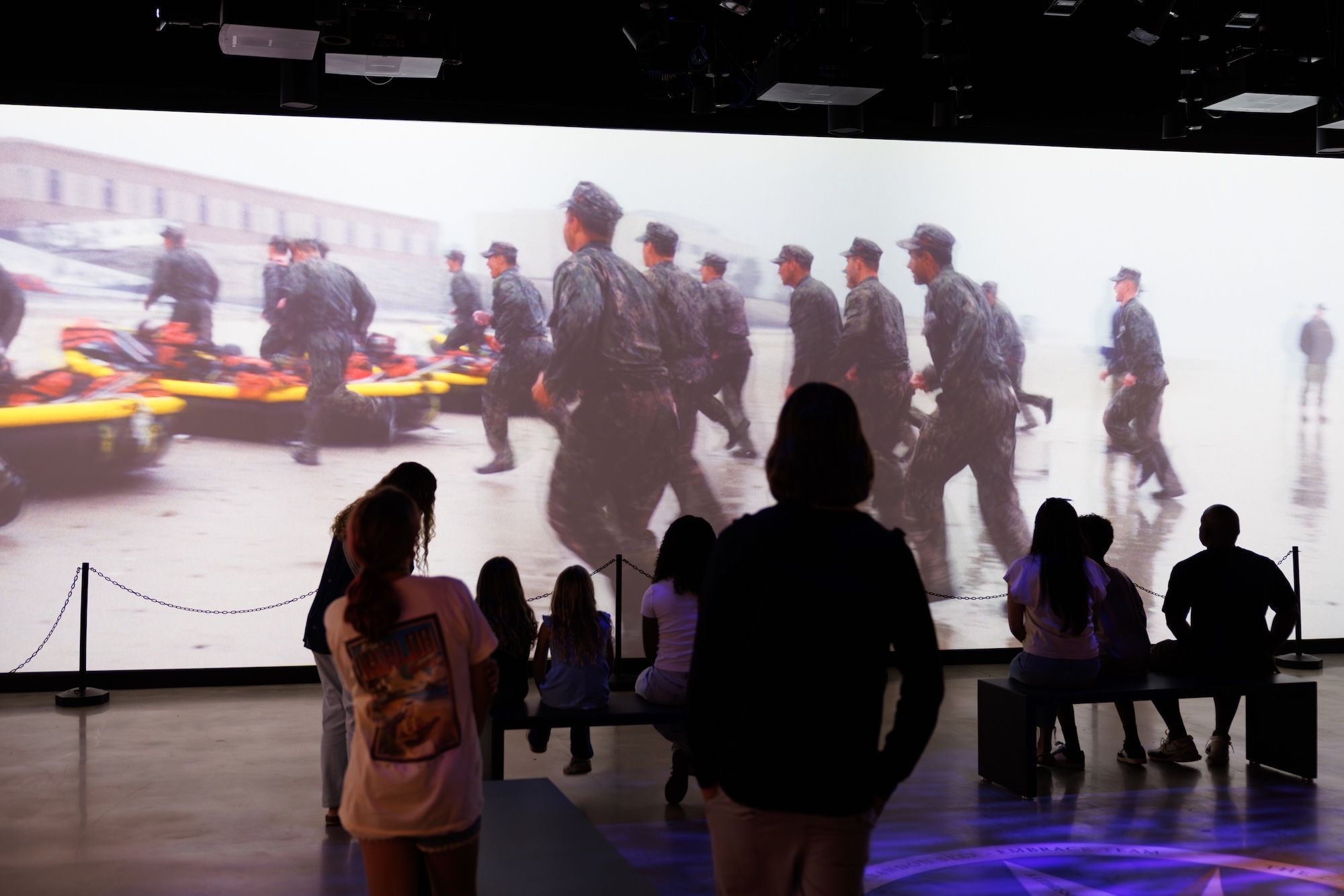 People watching a large screen showing soldiers in camouflage running beside inflatable rescue rafts on a wet surface in an indoor exhibit.
