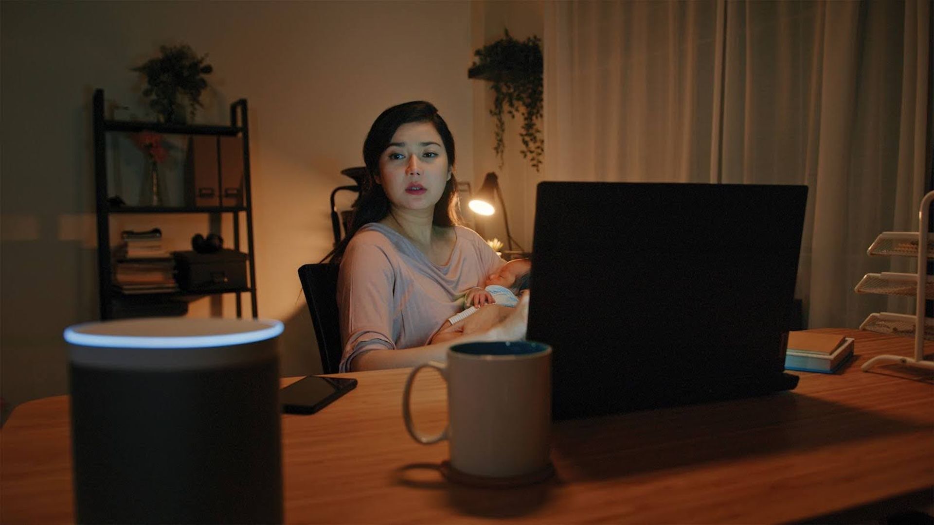 Woman working late at home on a laptop while holding a sleeping baby, with a smart speaker and coffee mug on the desk.