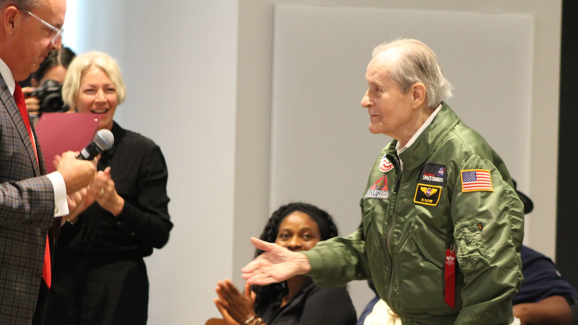 Elderly man in green pilot jacket with patches extends hand to a man in plaid suit and red tie holding a microphone, while seated people applaud in a bright room.
