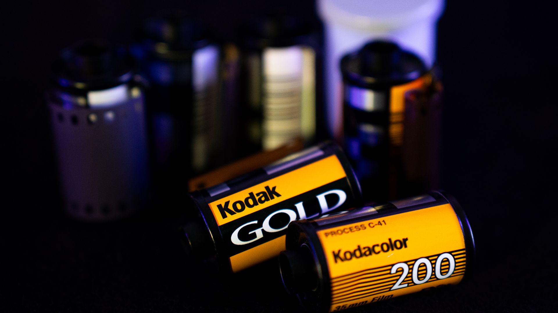 Close-up of Kodak 35mm film rolls, including Kodak Gold and Kodacolor 200, with black, yellow, and orange labels on a dark background.
