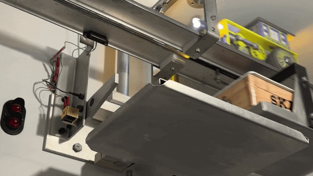 Industrial conveyor system with a grey metal frame and an inclined tray. A wooden crate and yellow-green packaged items sit on a shelf above, with exposed wiring and a red warning light.