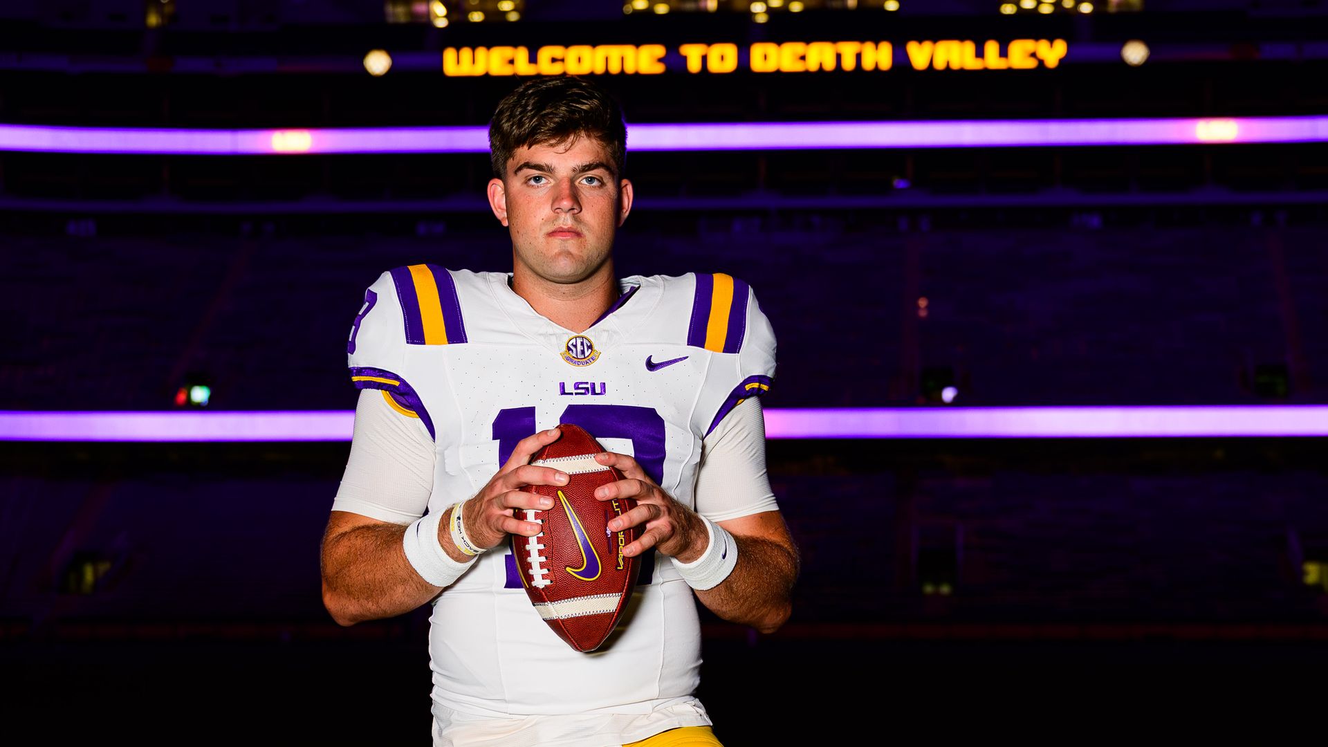 Football player in white, purple, and yellow LSU uniform holding a football, standing on green field at night with one foot on a yellow LSU helmet. "WELCOME TO DEATH VALLEY" sign lit in background.