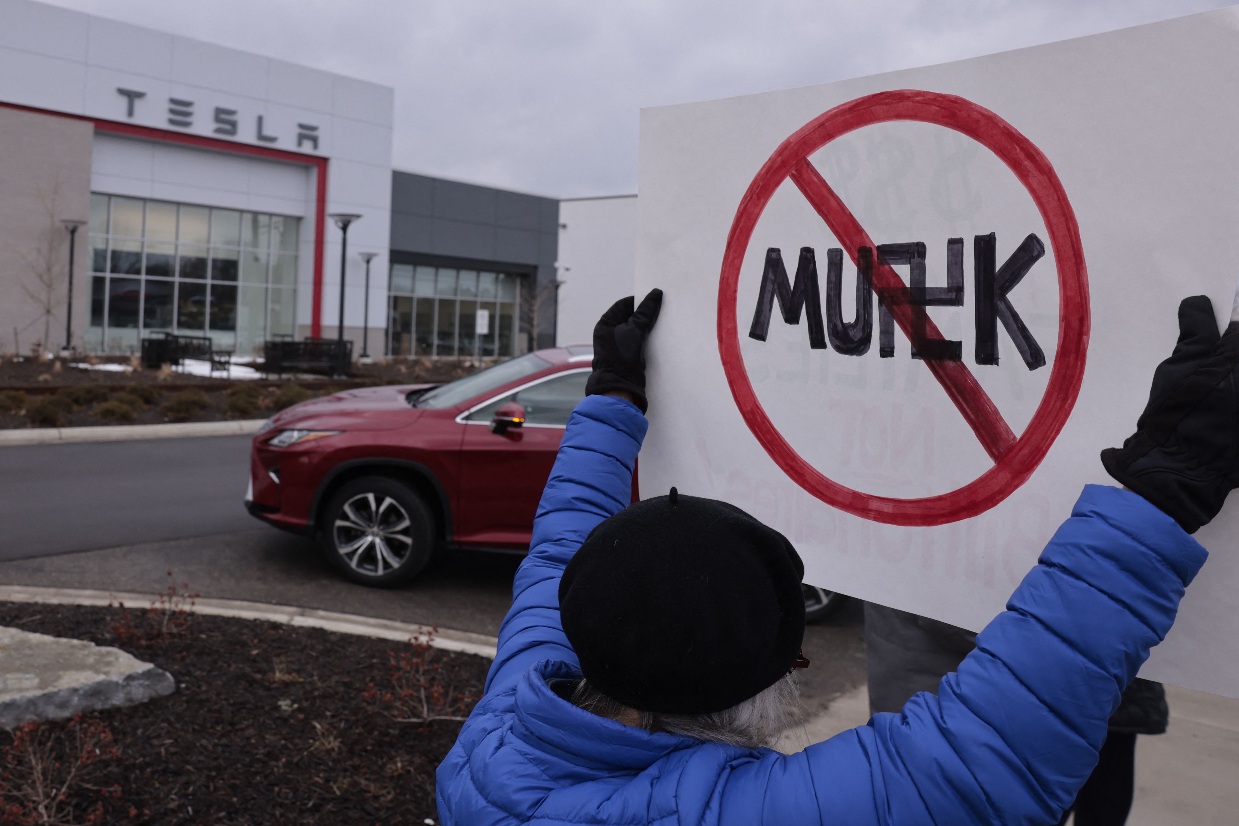 A person holds a sign that reads MUSK with an X over it and the S in his name is replaced for a Swastika