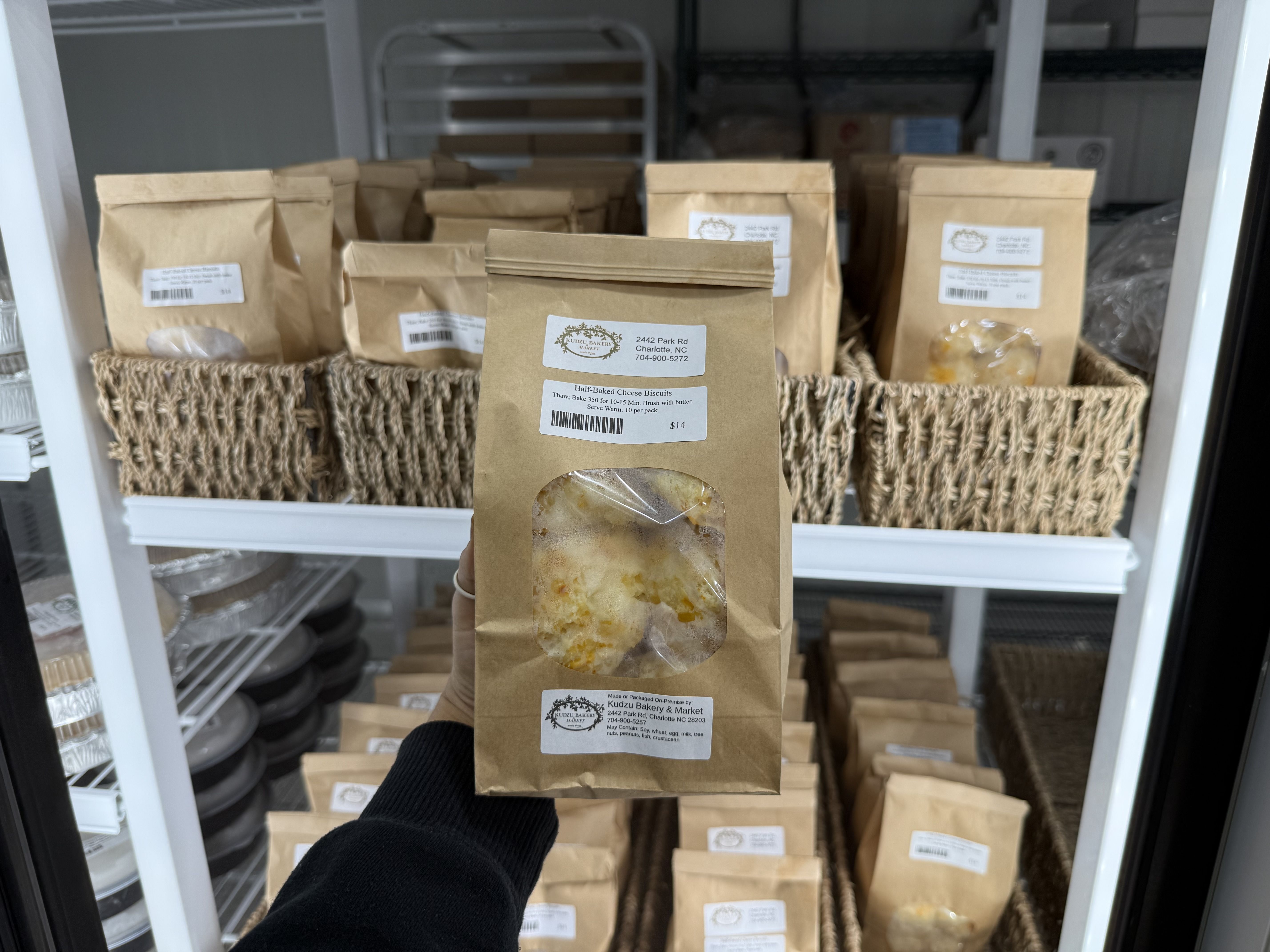 Hand holding a brown paper bag of half-baked cheese biscuits with clear window, in front of shelves filled with similar bags stored in woven baskets inside a fridge.