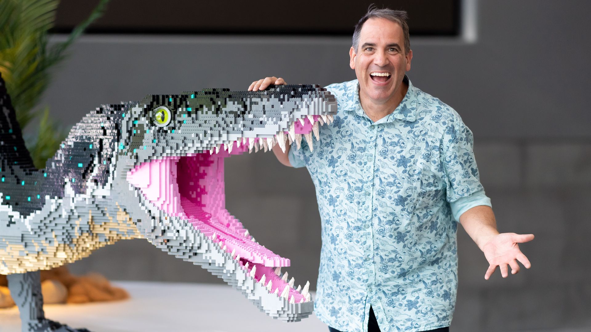 Smiling man in a light blue floral shirt stands beside a giant LEGO dinosaur made of gray, white, and pink bricks, resting a hand on its head; the dinosaur shows sharp teeth and a green eye.