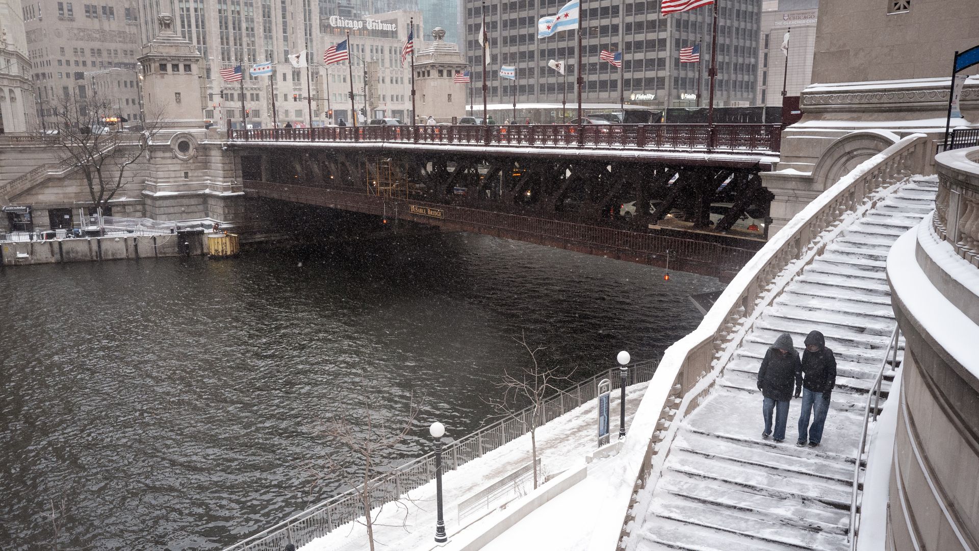 Photo of two people walking along a riverwalk in a city. 