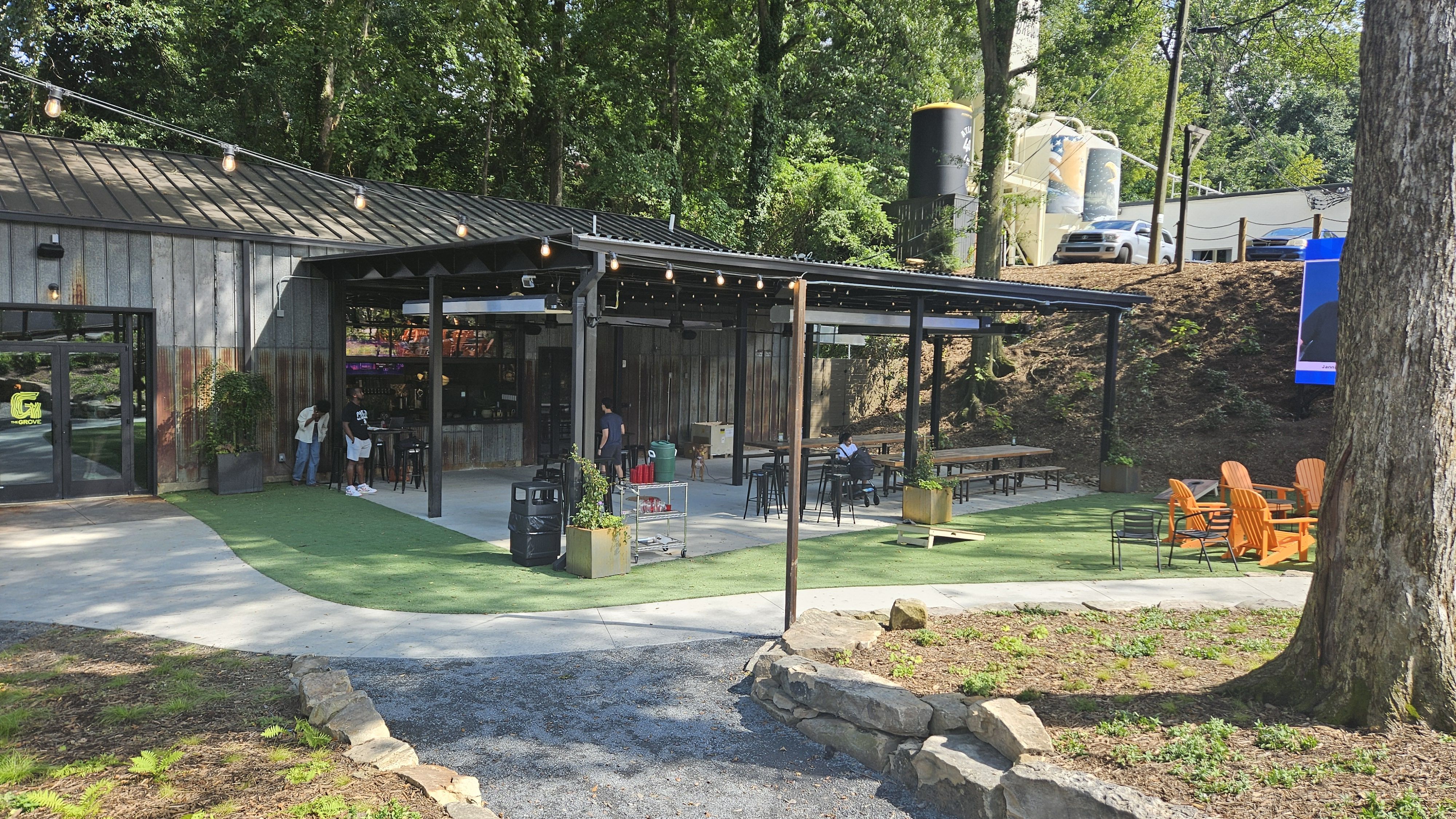 Outdoor brewery area with string lights, wooden tables, stools, and bright orange Adirondack chairs surrounded by trees and greenery under a metal roof structure.
