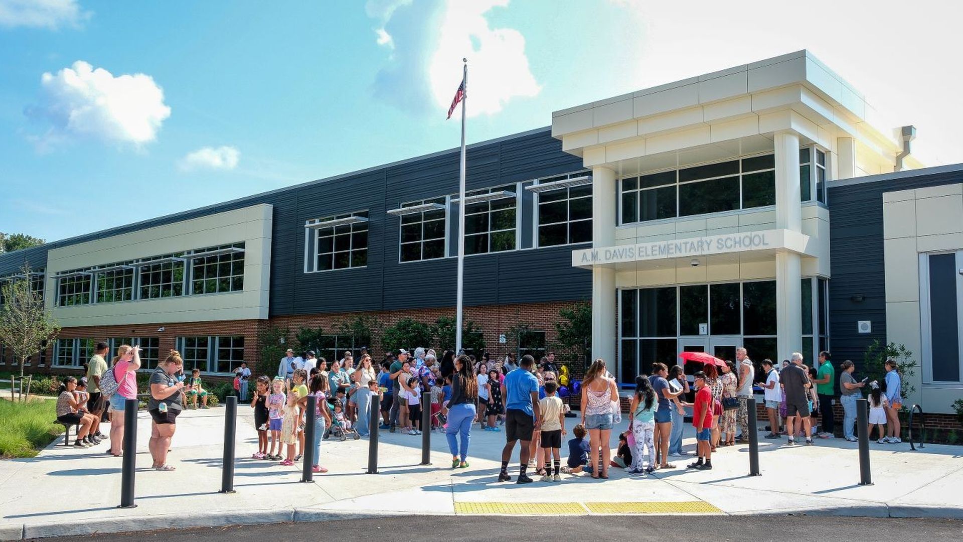 A group of adults and children gathered outside A.M. Davis Elementary School, a modern building with black, white, and brick exterior under a blue sky with scattered clouds.