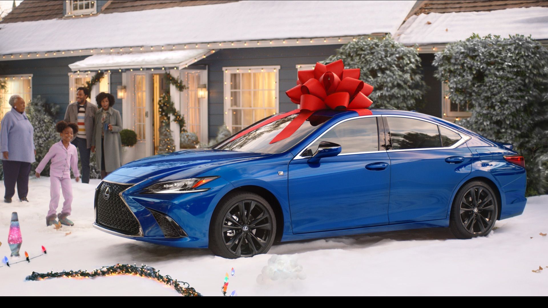 Blue Lexus sedan with a large red bow on top parked outside a house decorated with Christmas lights and garlands, with a happy family of five nearby in snowy yard.