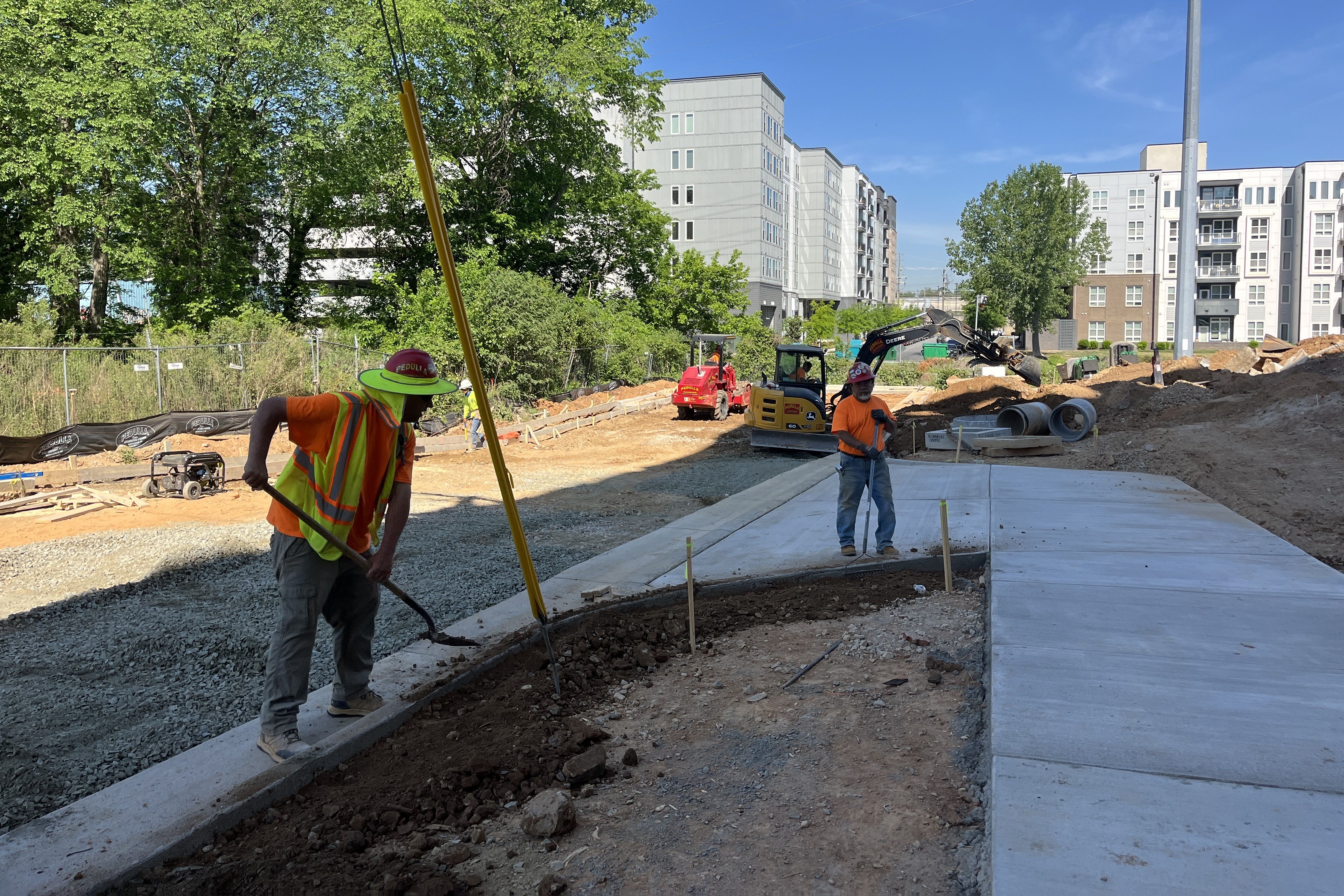 Construction site with workers in orange vests and helmets; dirt, gravel, and concrete along a new sidewalk. Modern multi-story buildings in the background, trees, blue sky, and machinery.