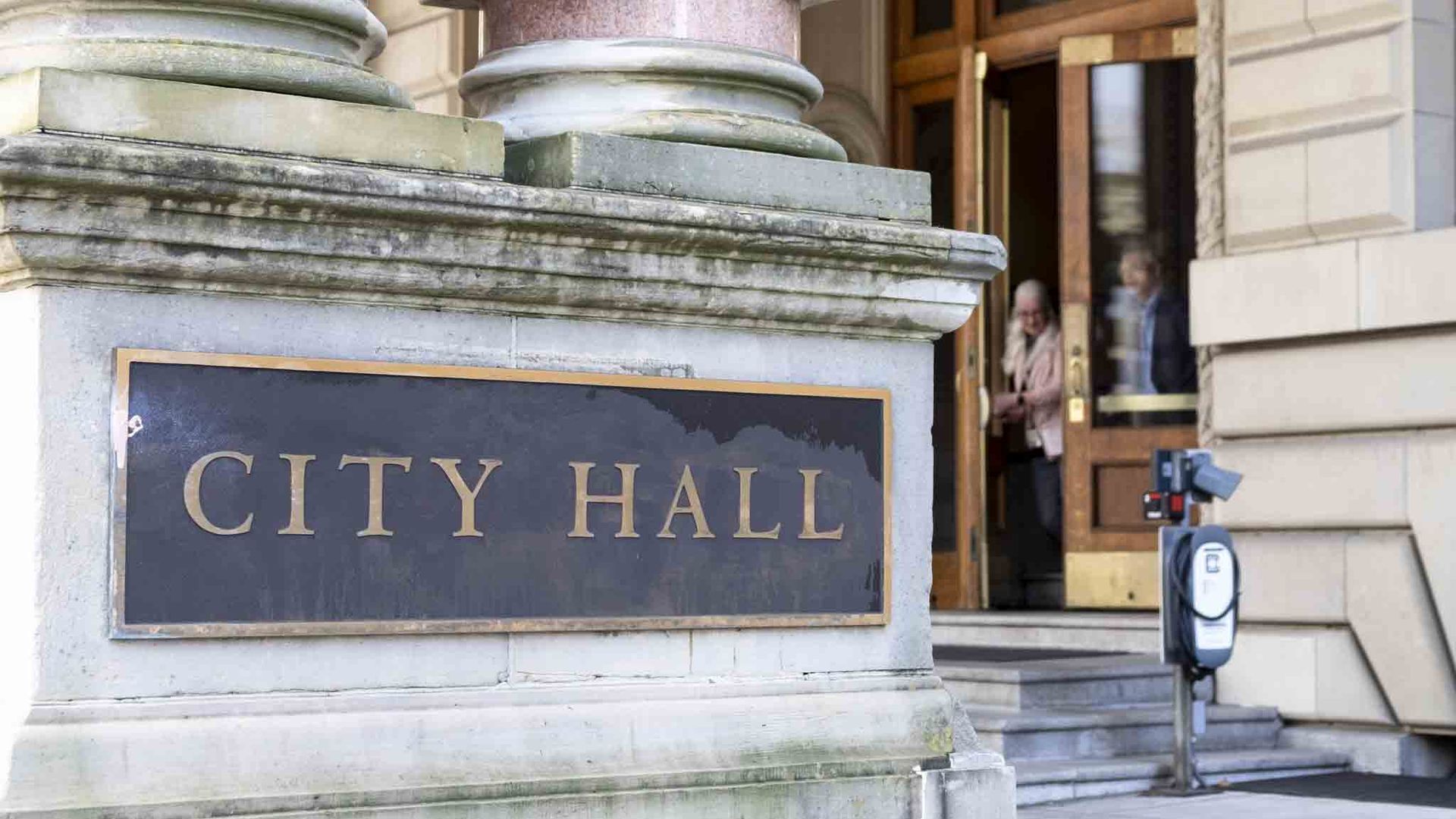 Close-up of a city hall façade with large stone columns and a dark plaque reading "CITY HALL" in gold letters. Open wooden doors reveal the entrance and a few people nearby.